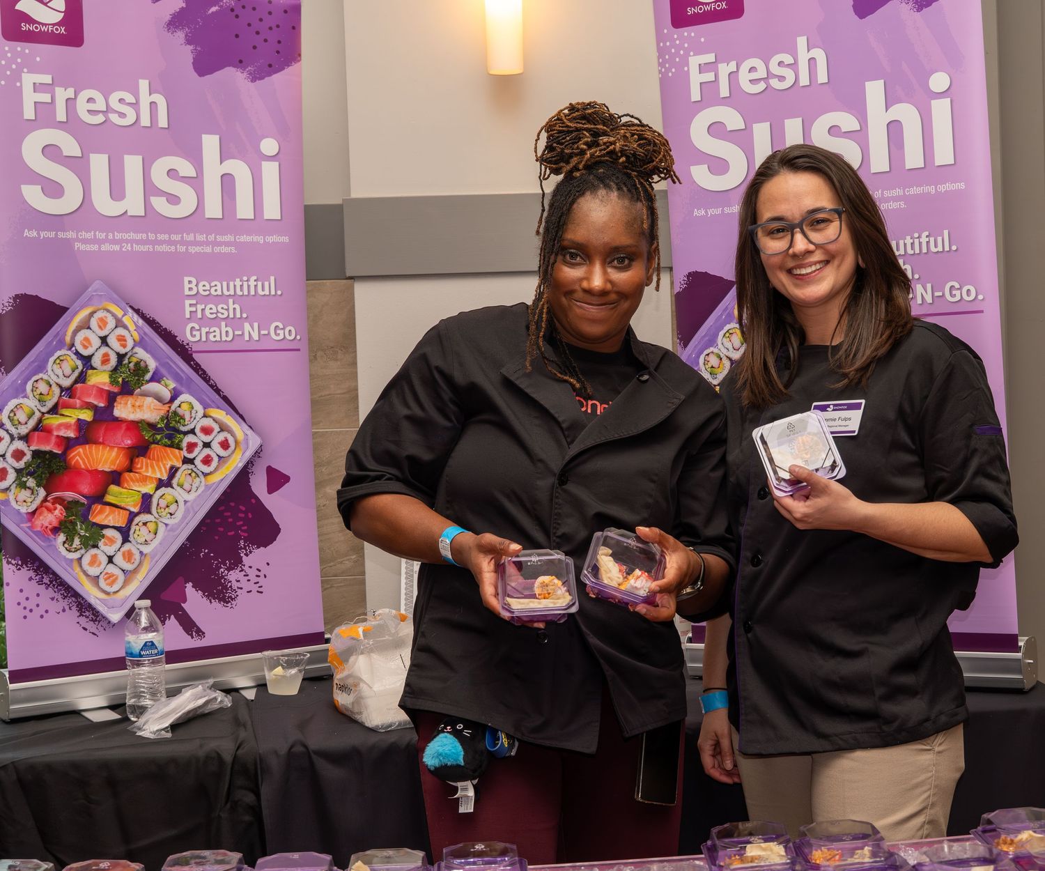 Two women smile at Fresh Sushi grab-and-go booth displaying colorful sushi platters at food event.