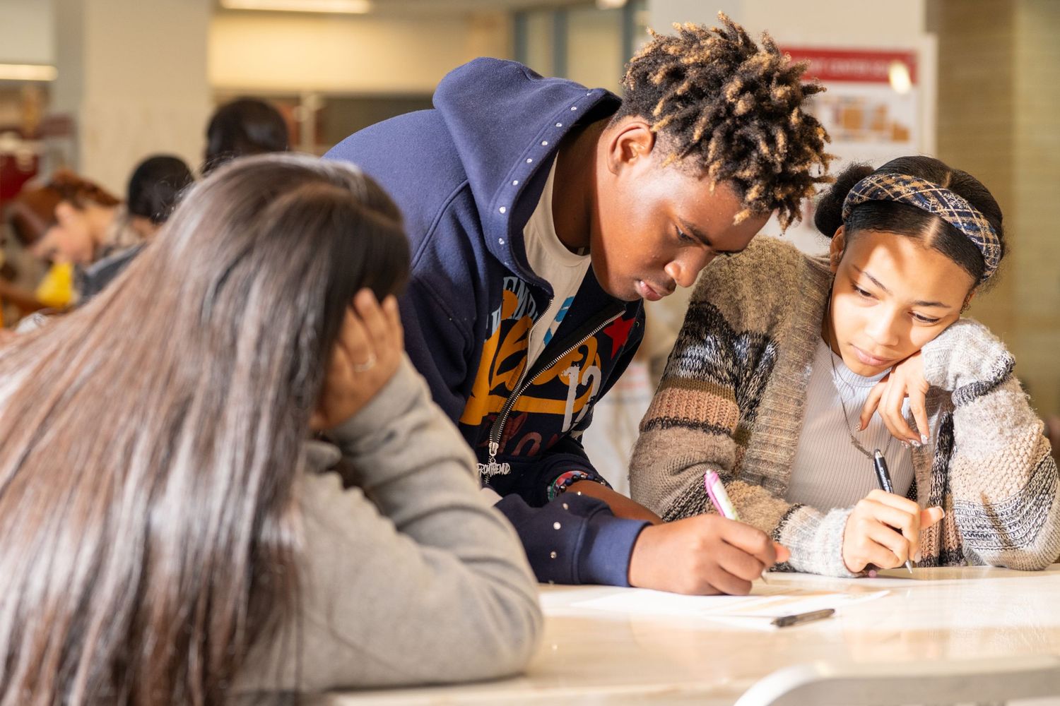 Three students collaborate closely on a writing assignment at a table in a busy school setting.