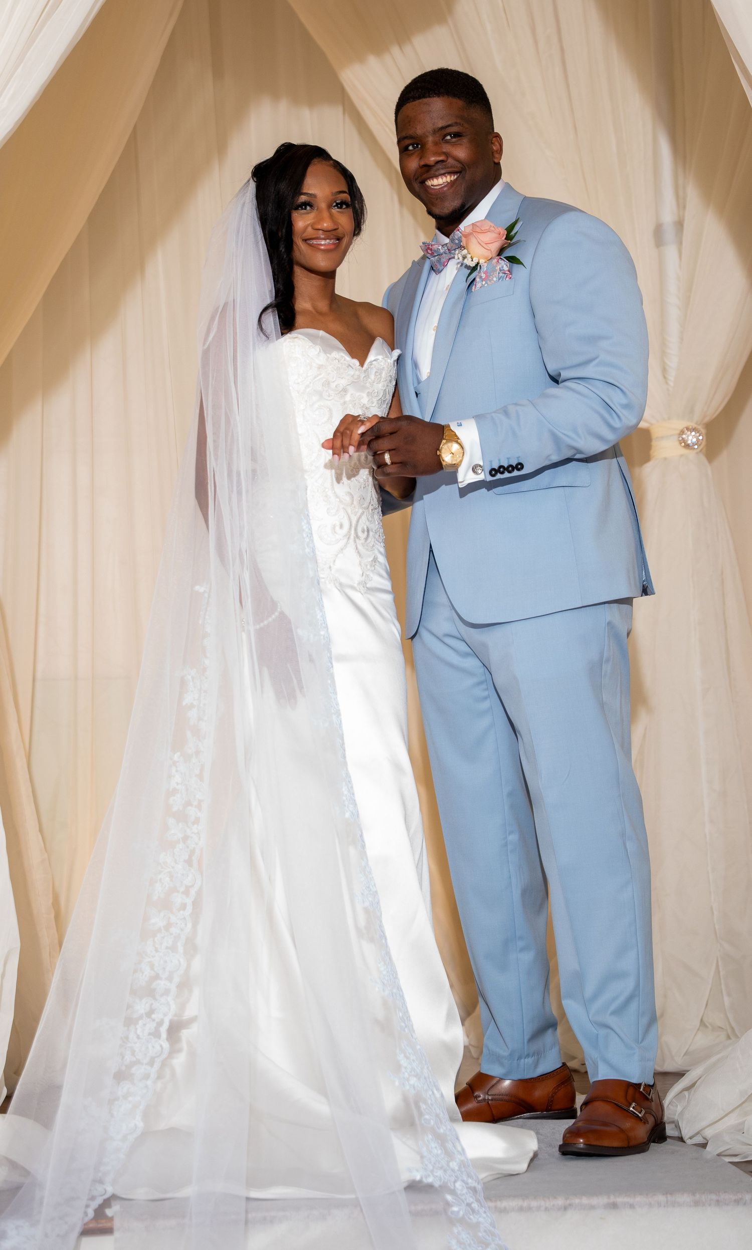 Newlywed couple posing elegantly, groom in light blue suit and bride in white wedding gown with veil.