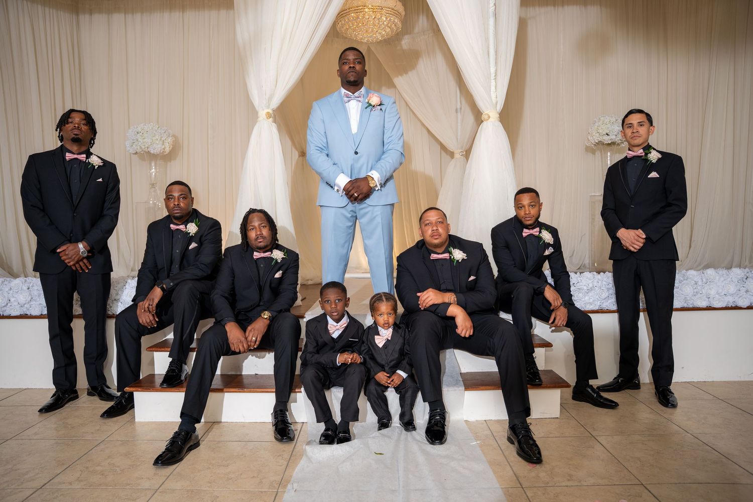 Groom in light blue suit stands with groomsmen in black tuxedos at elegant wedding venue.