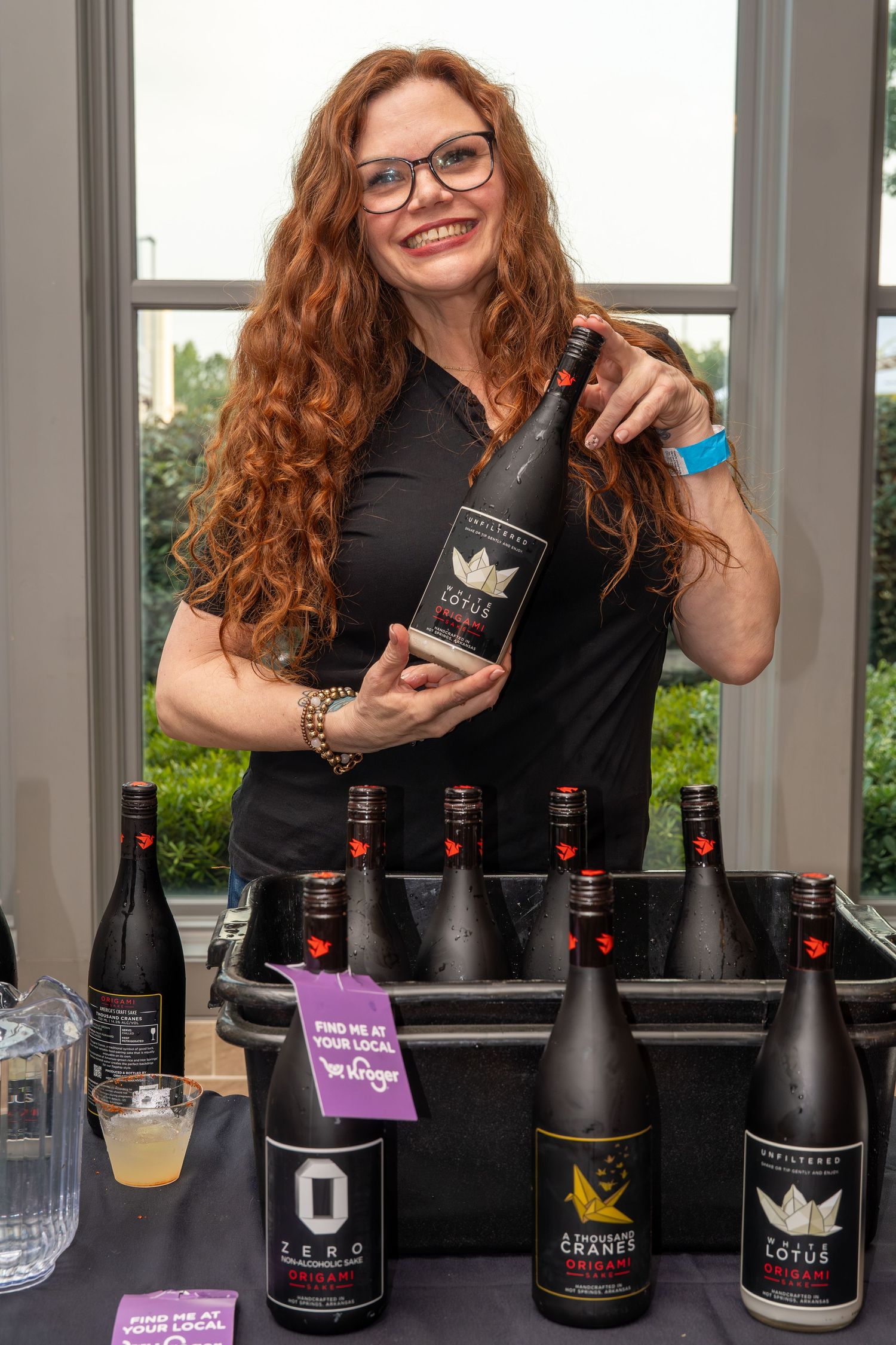 Smiling redhead woman holds Lotus wine bottle at vendor table displaying Mediterranean wine collection.