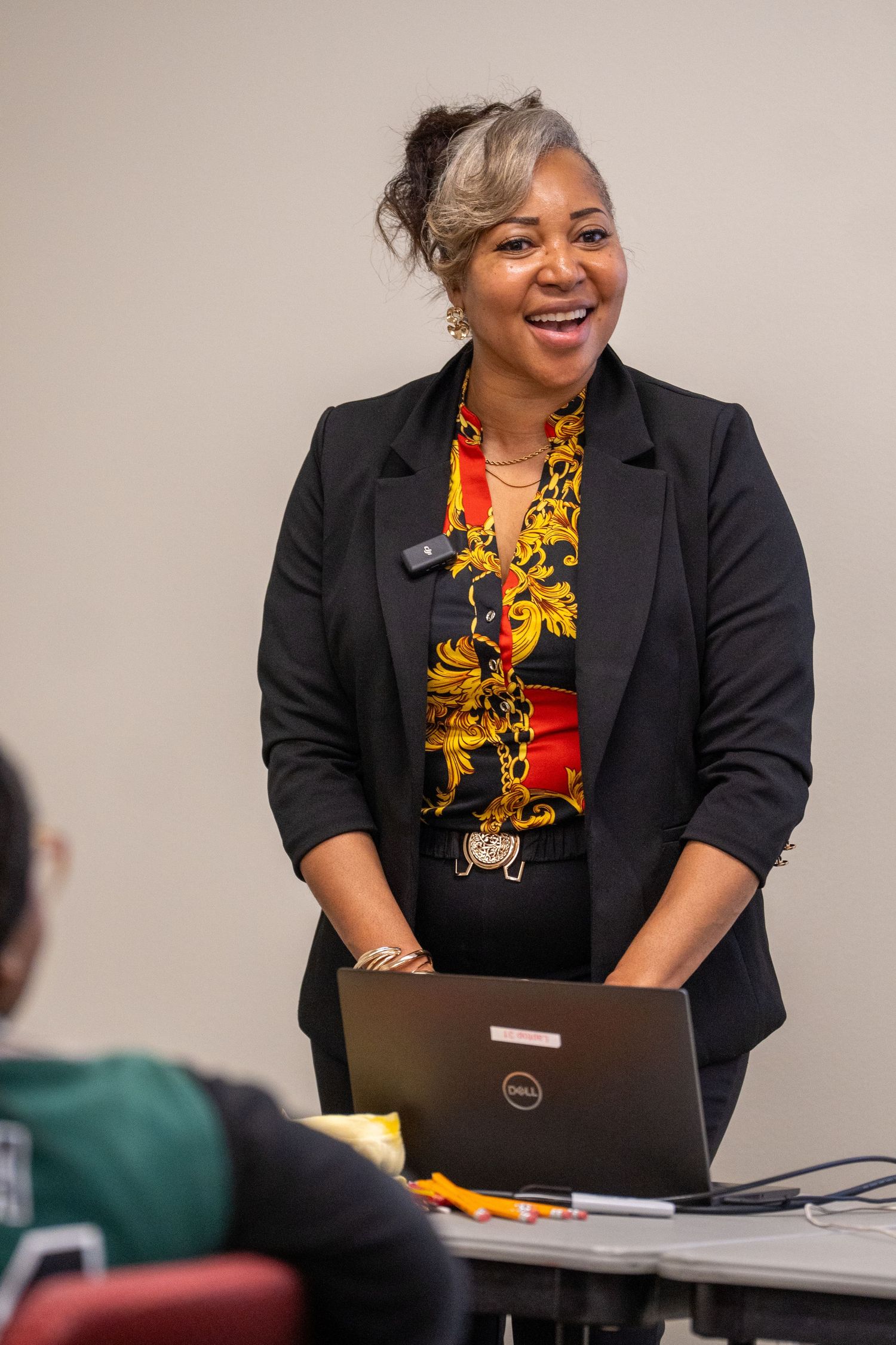 Smiling woman in black blazer and gold patterned blouse presents beside a Dell laptop in classroom.