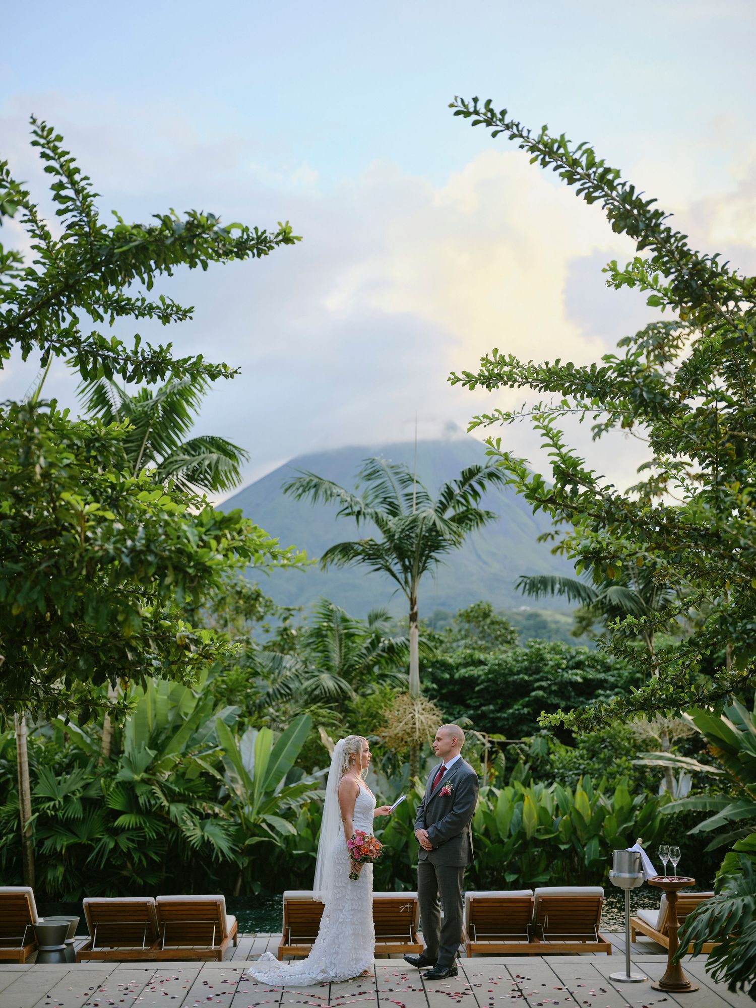 Bride and groom exchanging vows on a terrace framed by tropical trees with a volcano backdrop.