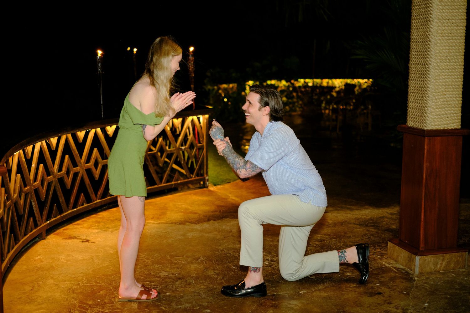 Man proposes on one knee to surprised woman in green dress on illuminated bridge at night.