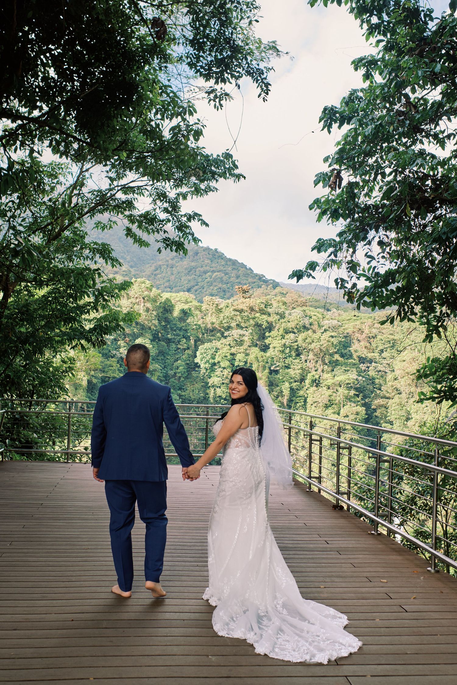 Bride and groom hold hands on a wooden deck overlooking lush green mountains on their wedding day.
