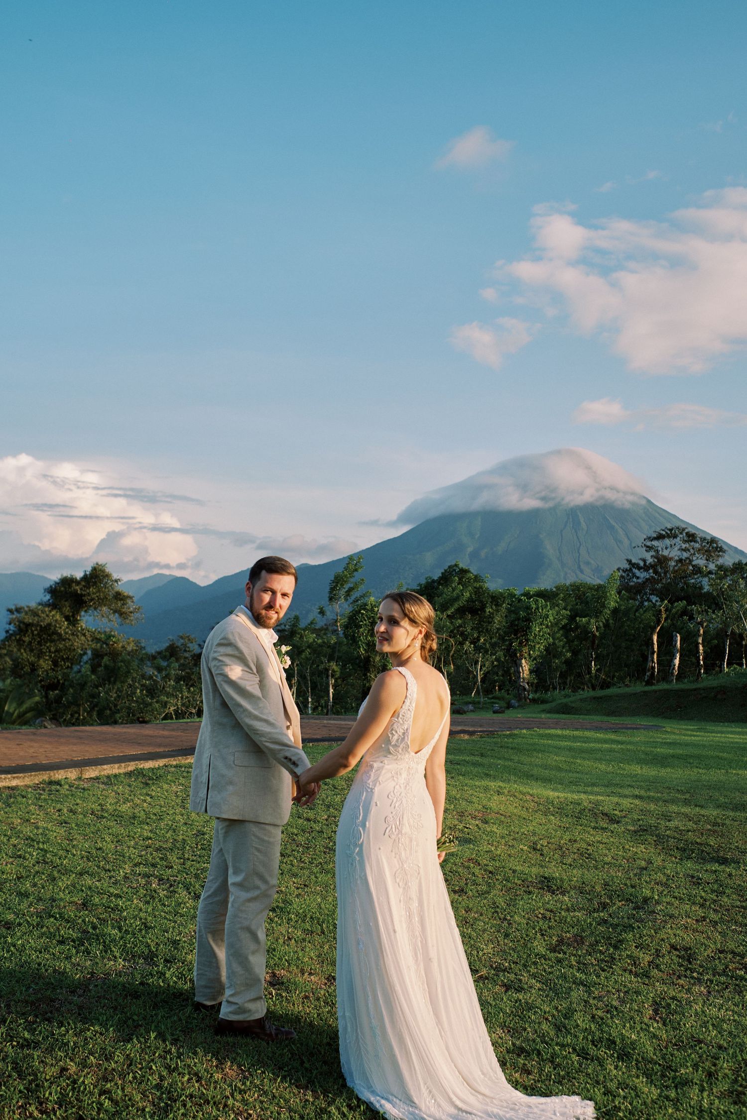 Bride and groom hold hands on a green lawn with a majestic volcano and blue sky in the background.