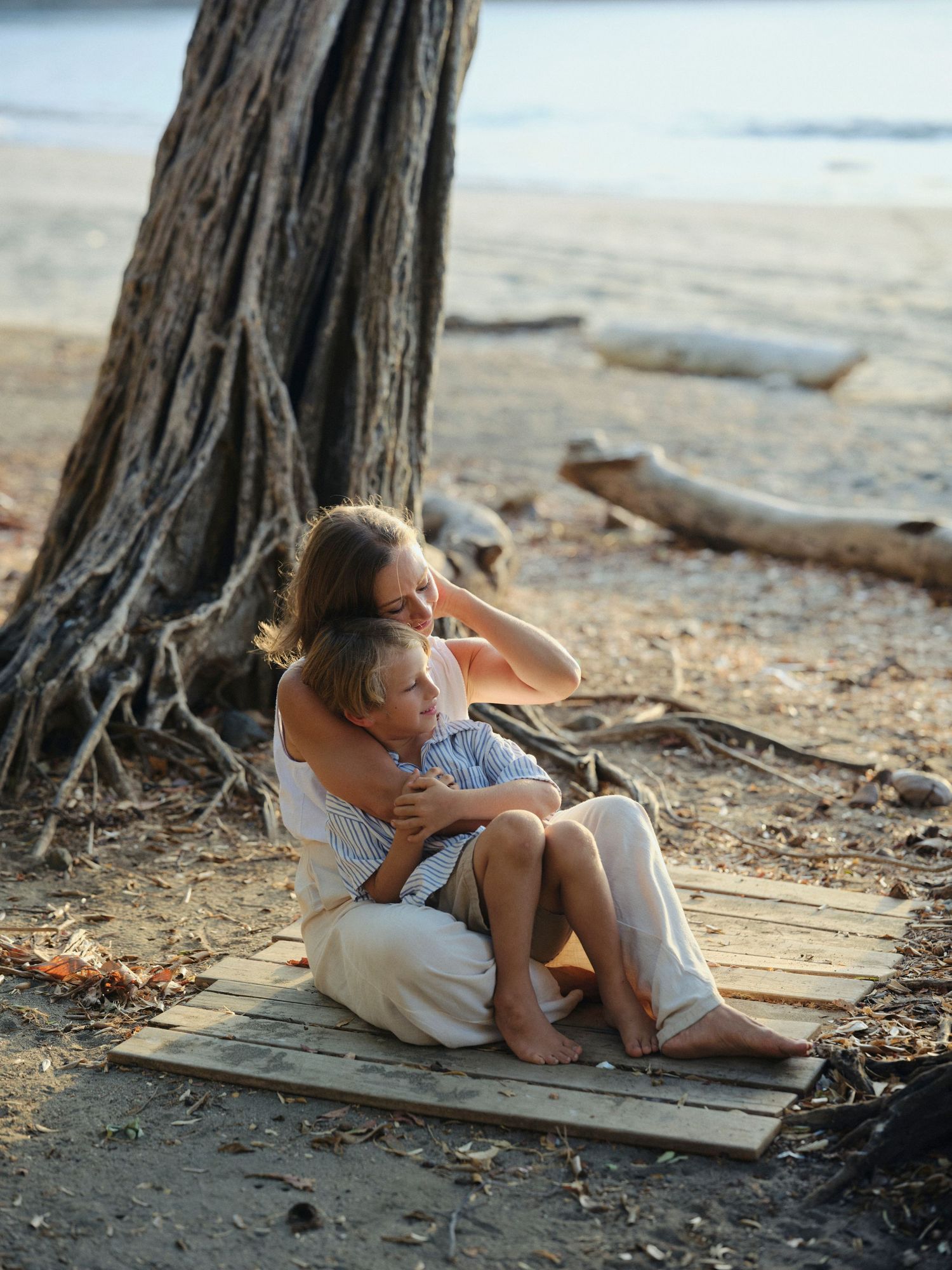 Mother and child sitting together under large tree roots on a peaceful tropical beach at golden hour.