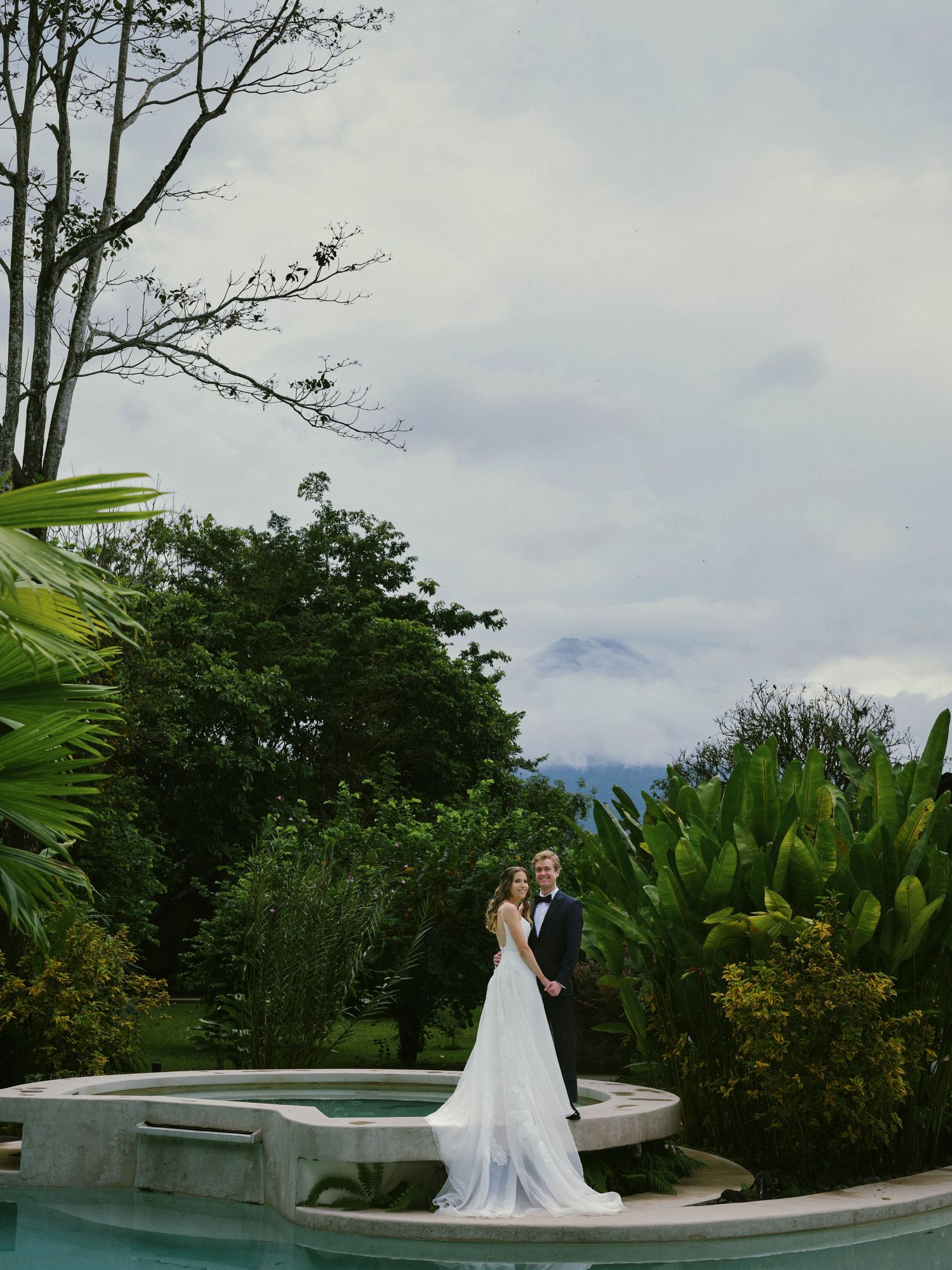 Bride and groom pose elegantly by a fountain surrounded by lush tropical gardens on their wedding day.