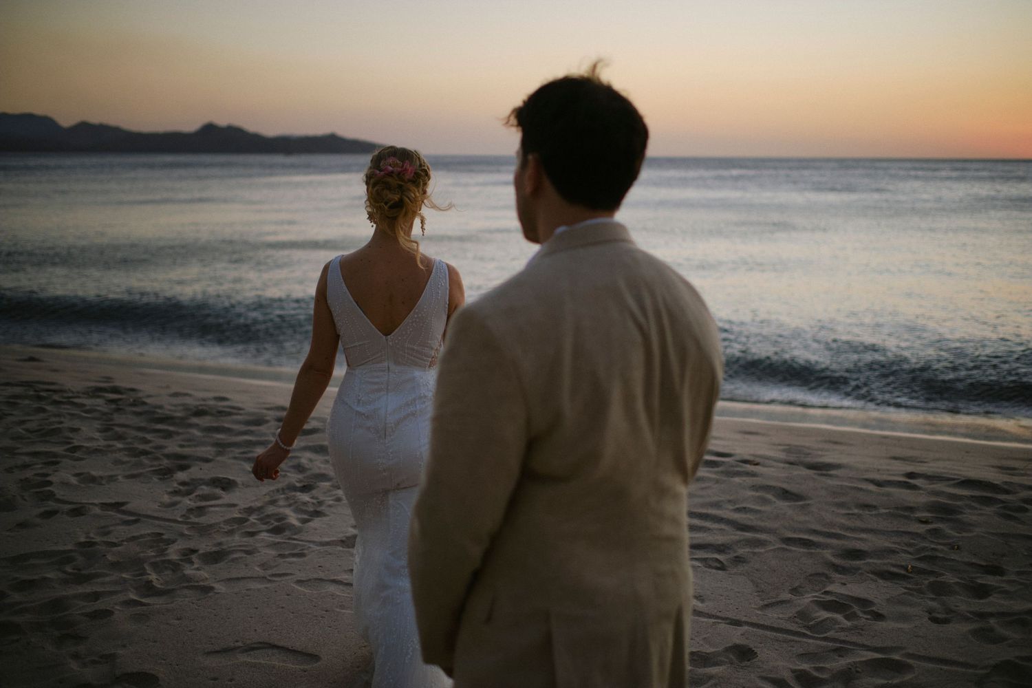 Bride and groom walk hand in hand along the beach at sunset after their wedding.