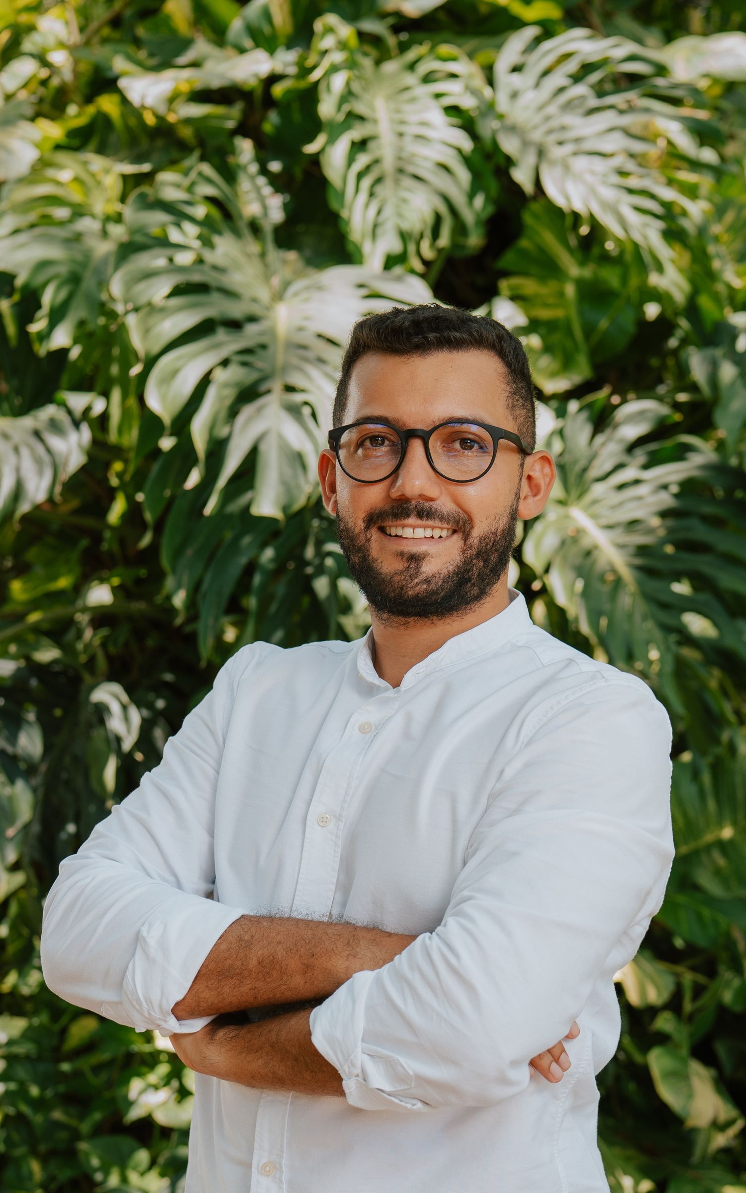 Smiling man with glasses and beard, arms crossed, wearing white shirt, standing in front of lush tropical green foliage.