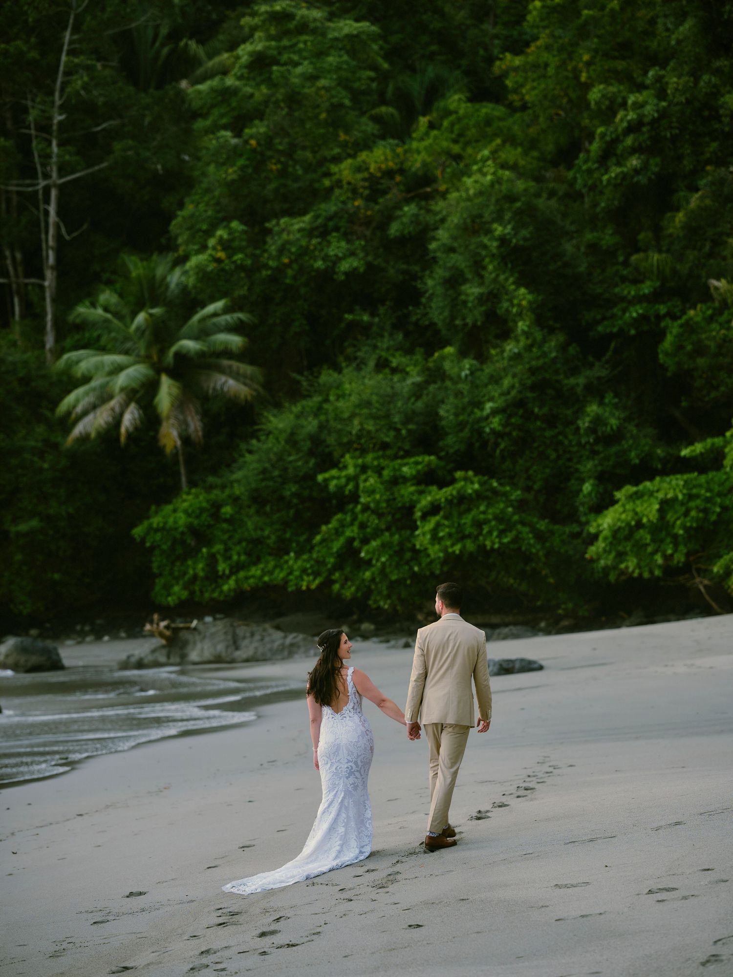 Bride and groom walking hand in hand along tropical beach with lush jungle backdrop.