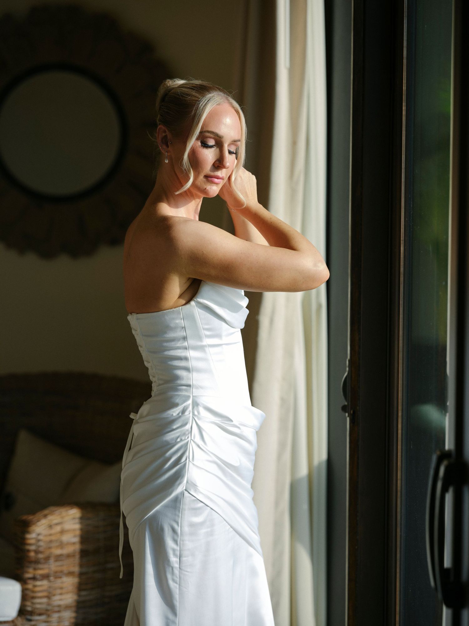 Elegant bride in strapless white ruched wedding gown adjusts earring by sunlit glass door.
