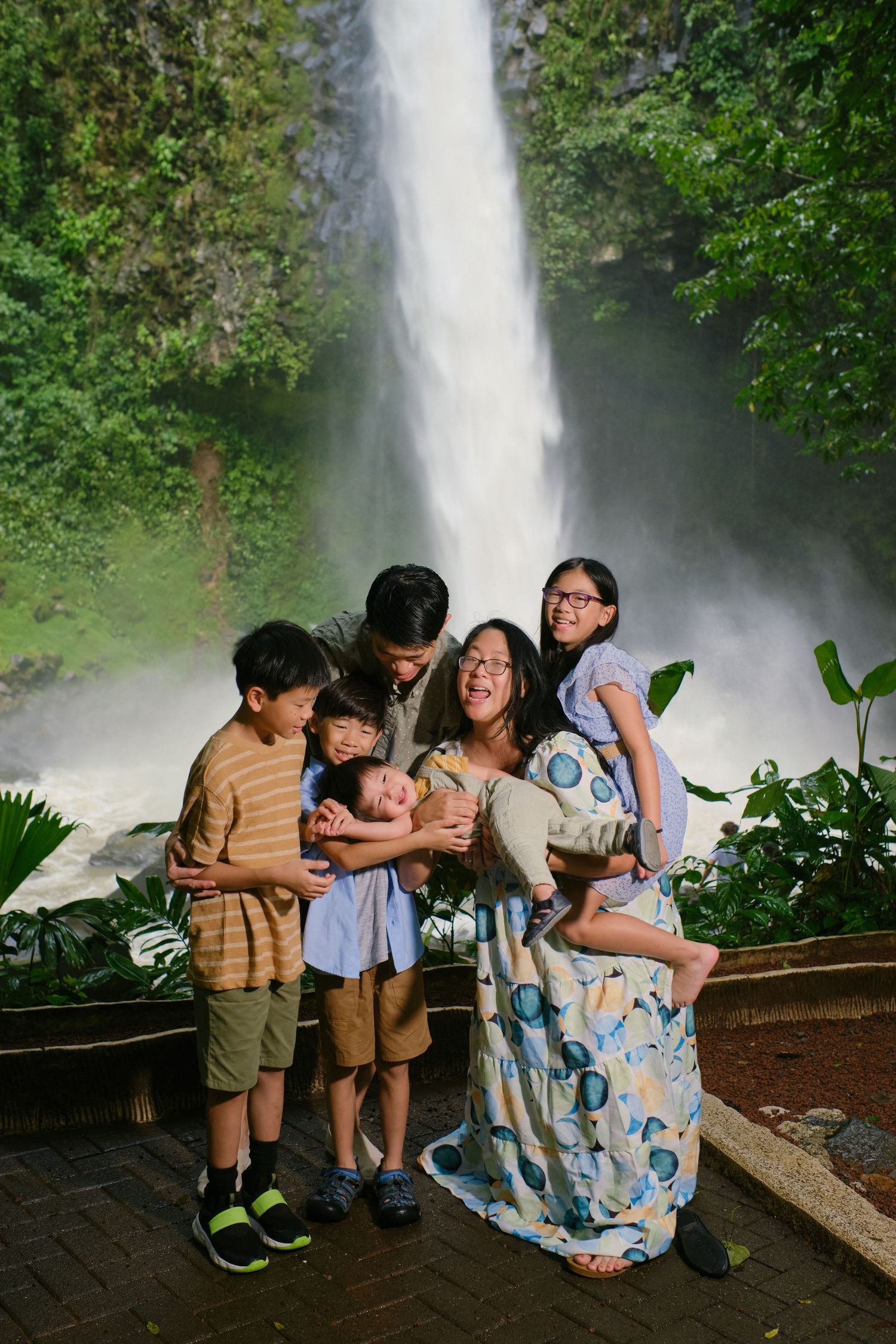 Happy family of six posing and laughing in front of a stunning tall waterfall in a tropical setting.