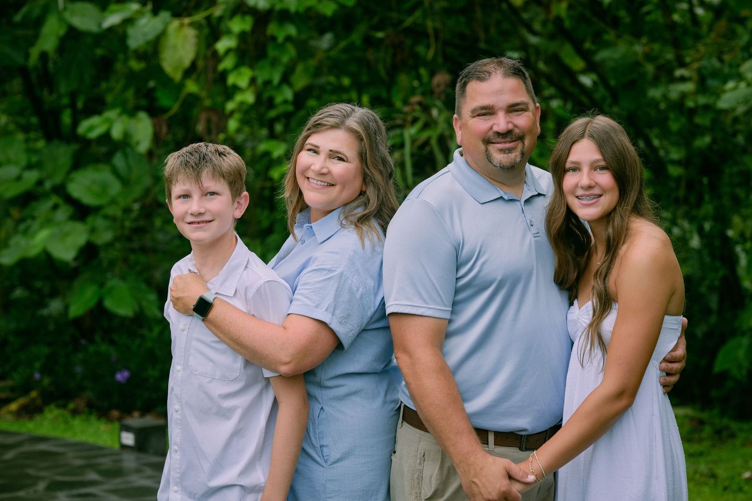 Happy family of four in coordinating blue outfits smiling together in front of lush tropical greenery.
