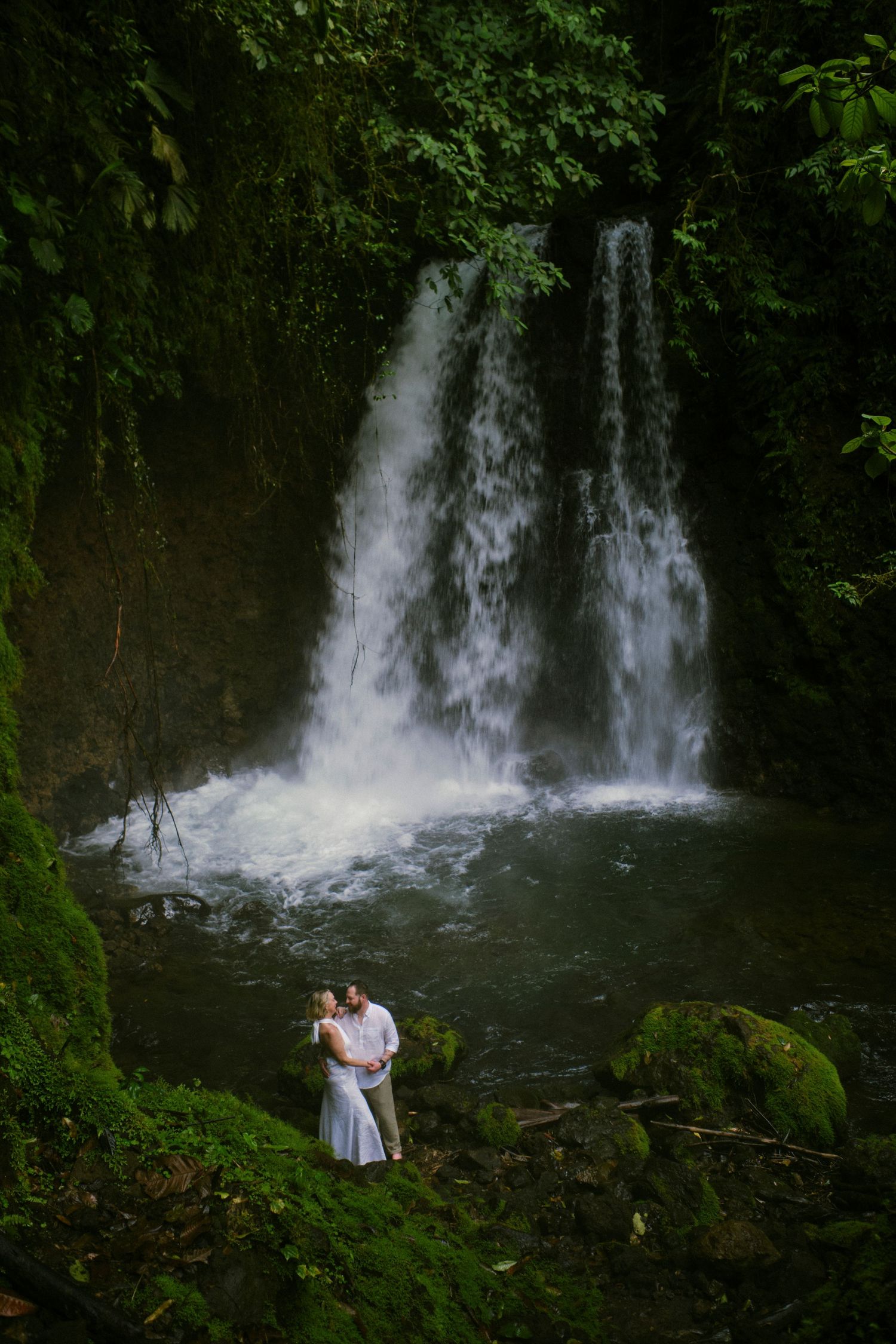 Couple in white embracing romantically in front of a stunning double waterfall in a lush green forest.