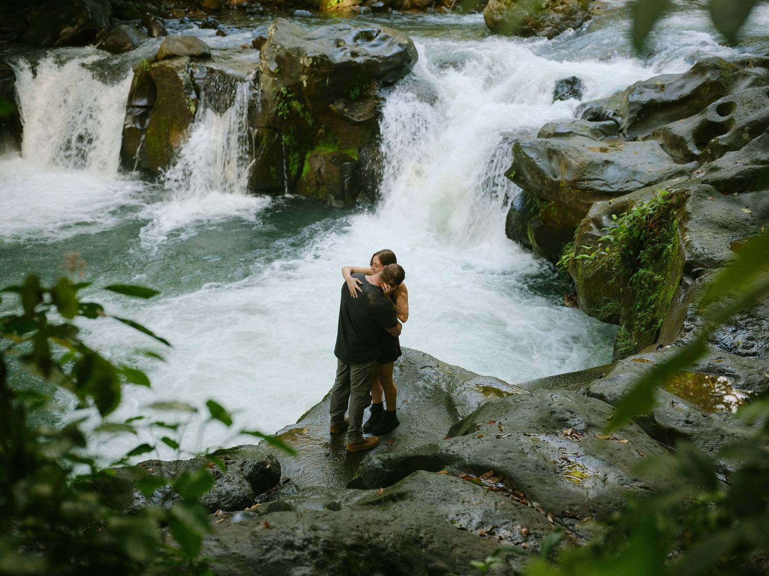 Couple embraces on mossy rocks surrounded by cascading waterfalls in a lush green jungle.