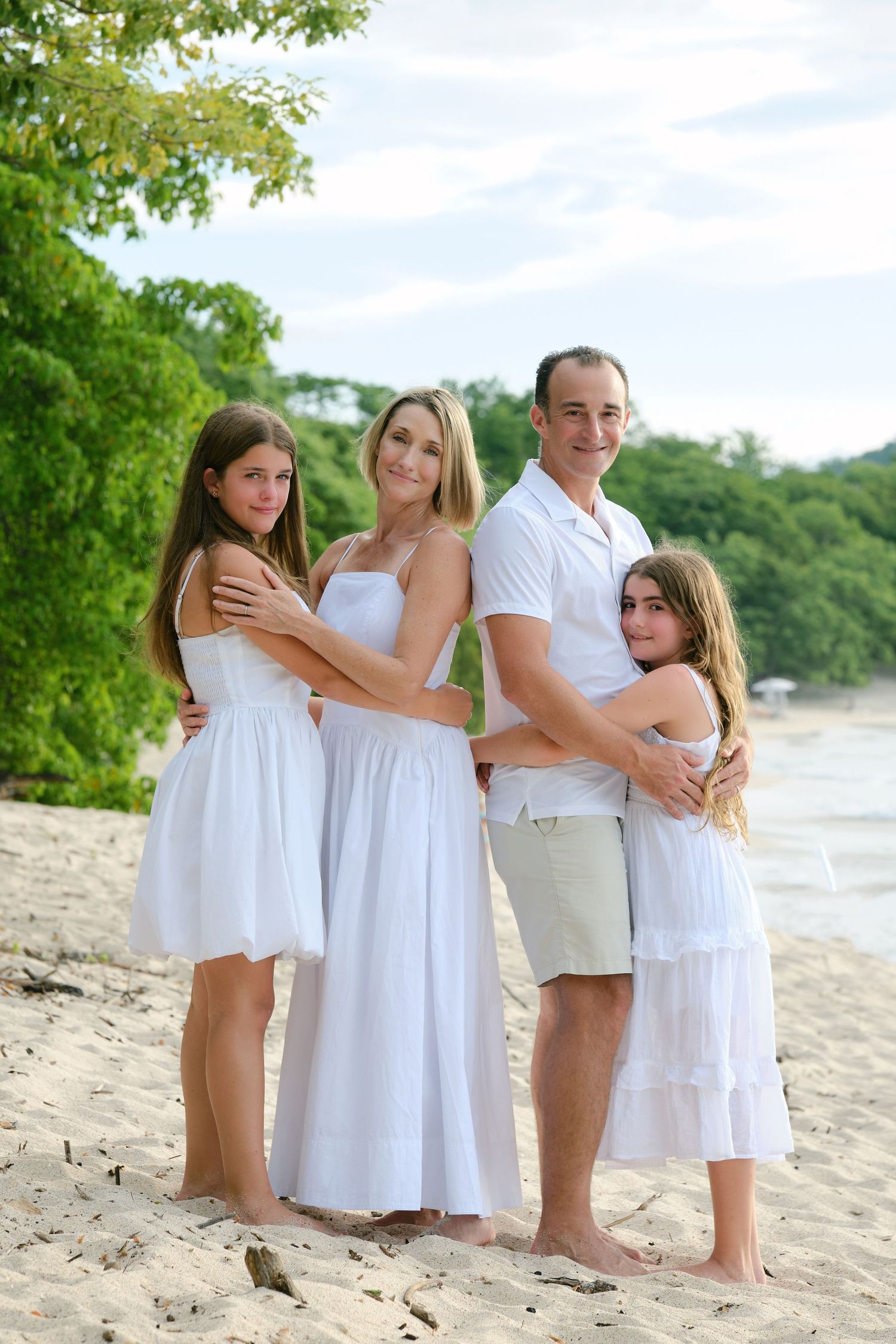 Family of four in matching white outfits embracing on a tropical beach with lush green trees behind them.