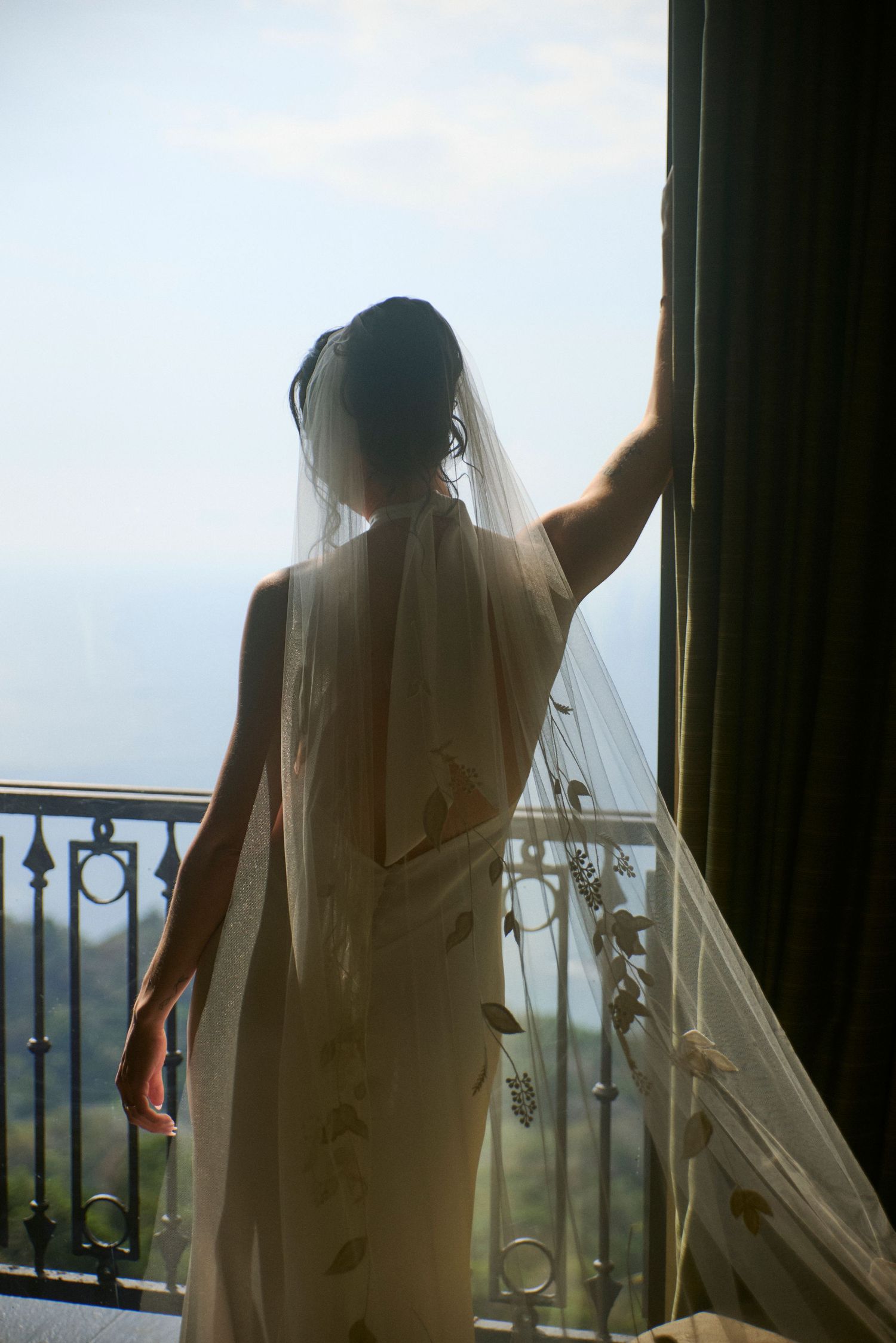 Bride in elegant halter gown and floral veil stands backlit by a balcony with ocean views.