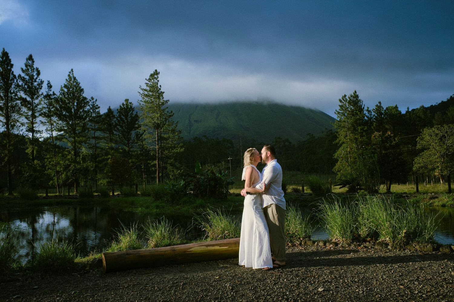 Bride and groom standing by a reflective pond with misty mountain and pine trees at dusk.