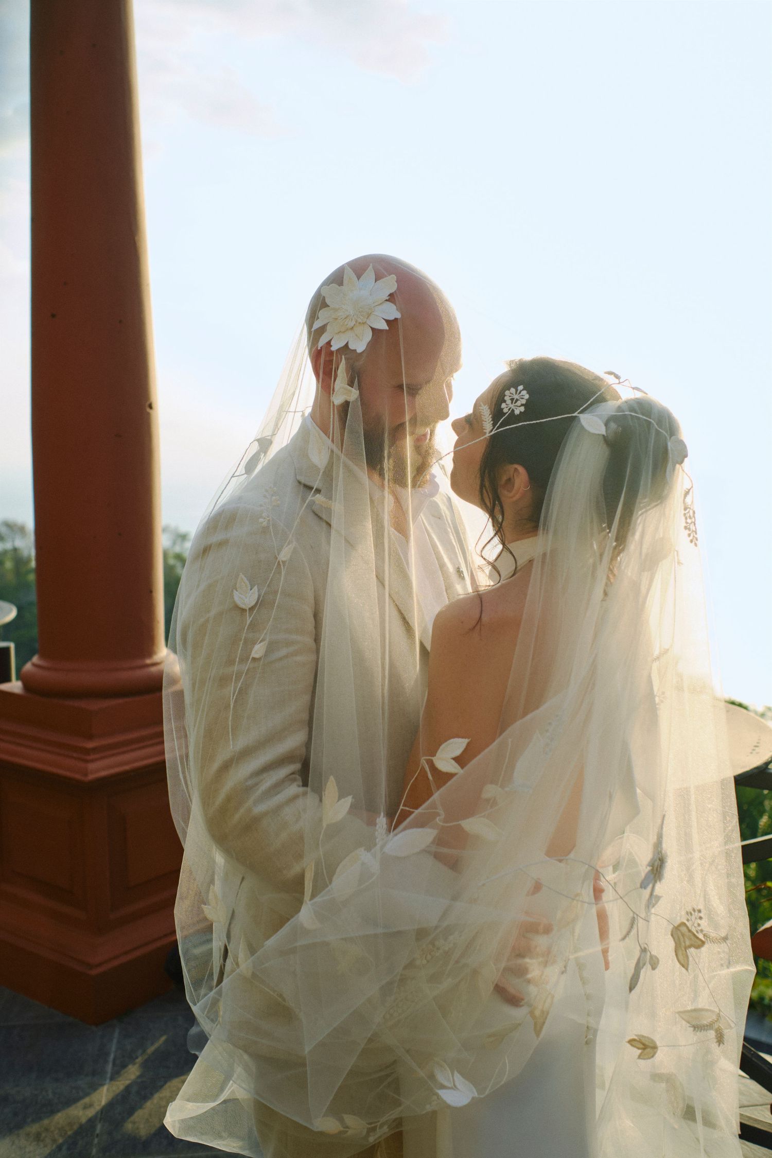 Bride and groom share an intimate moment under a floral veil in soft golden sunlight.