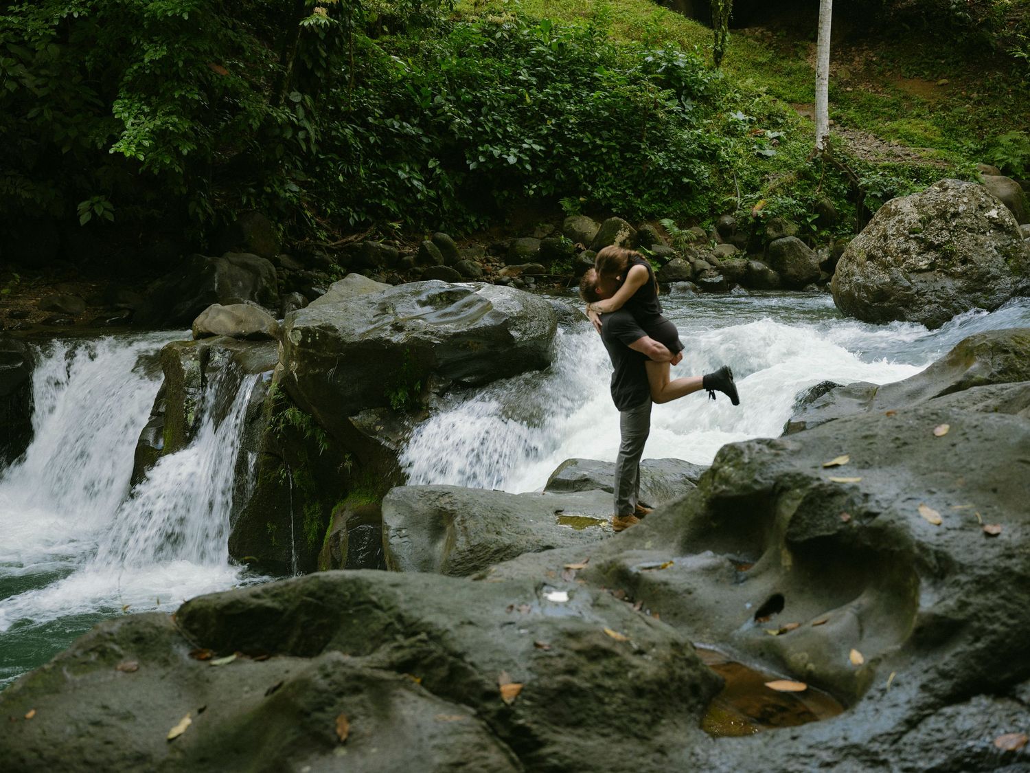 Woman joyfully jumps on rocks beside a cascading waterfall in a tropical jungle setting.