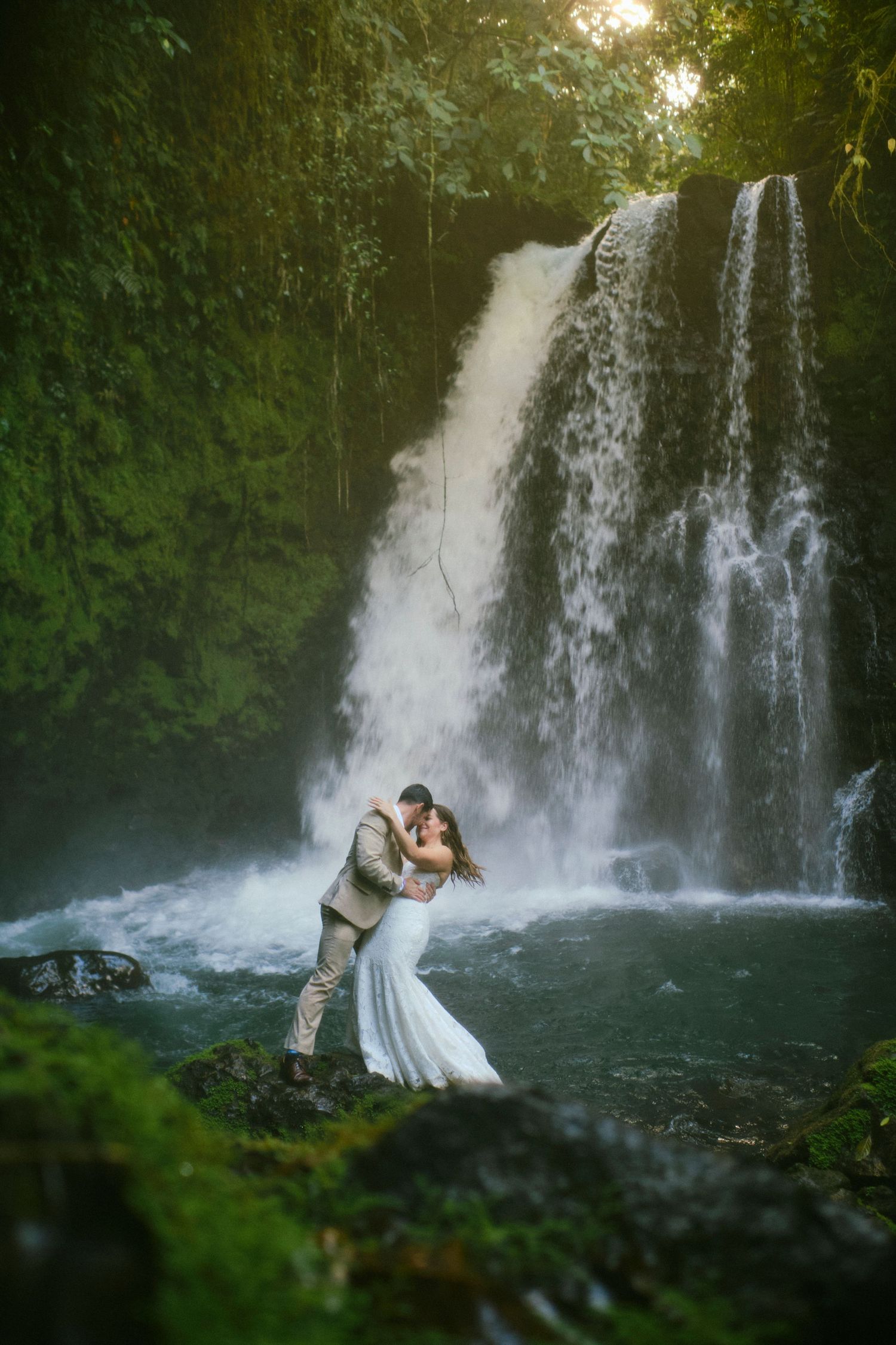 Bride and groom share a romantic kiss in front of a stunning lush waterfall surrounded by green tropical foliage.