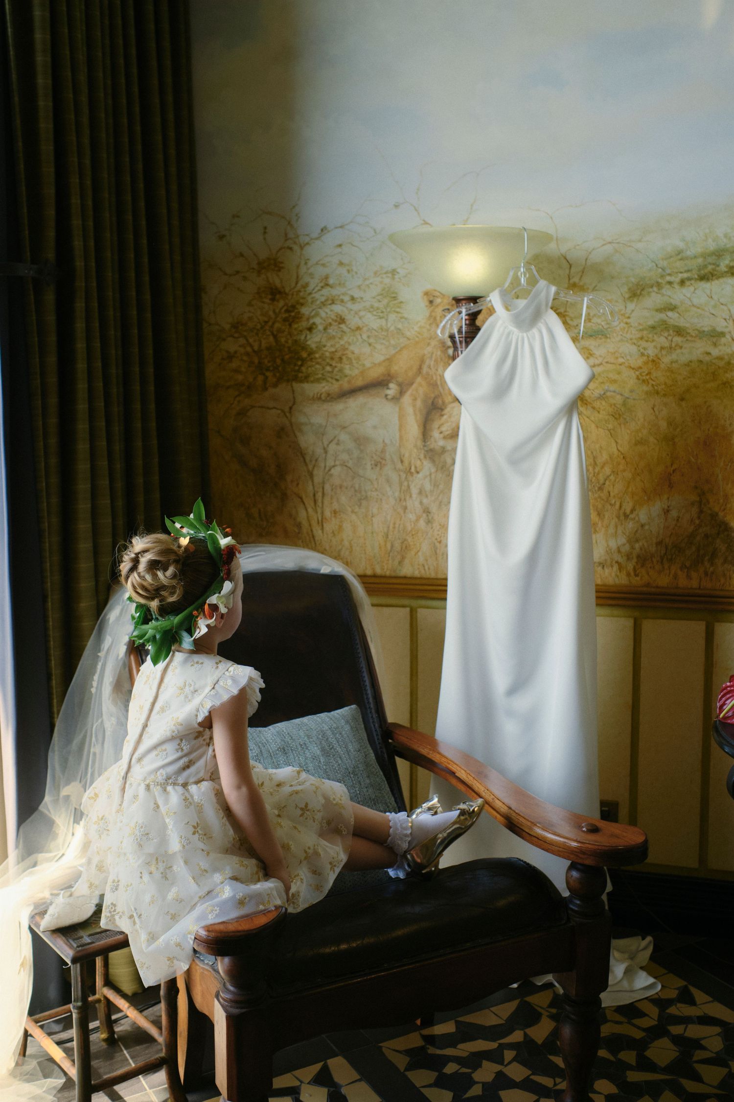 Flower girl in white dress admires a halter wedding gown hanging in a warmly lit safari room.