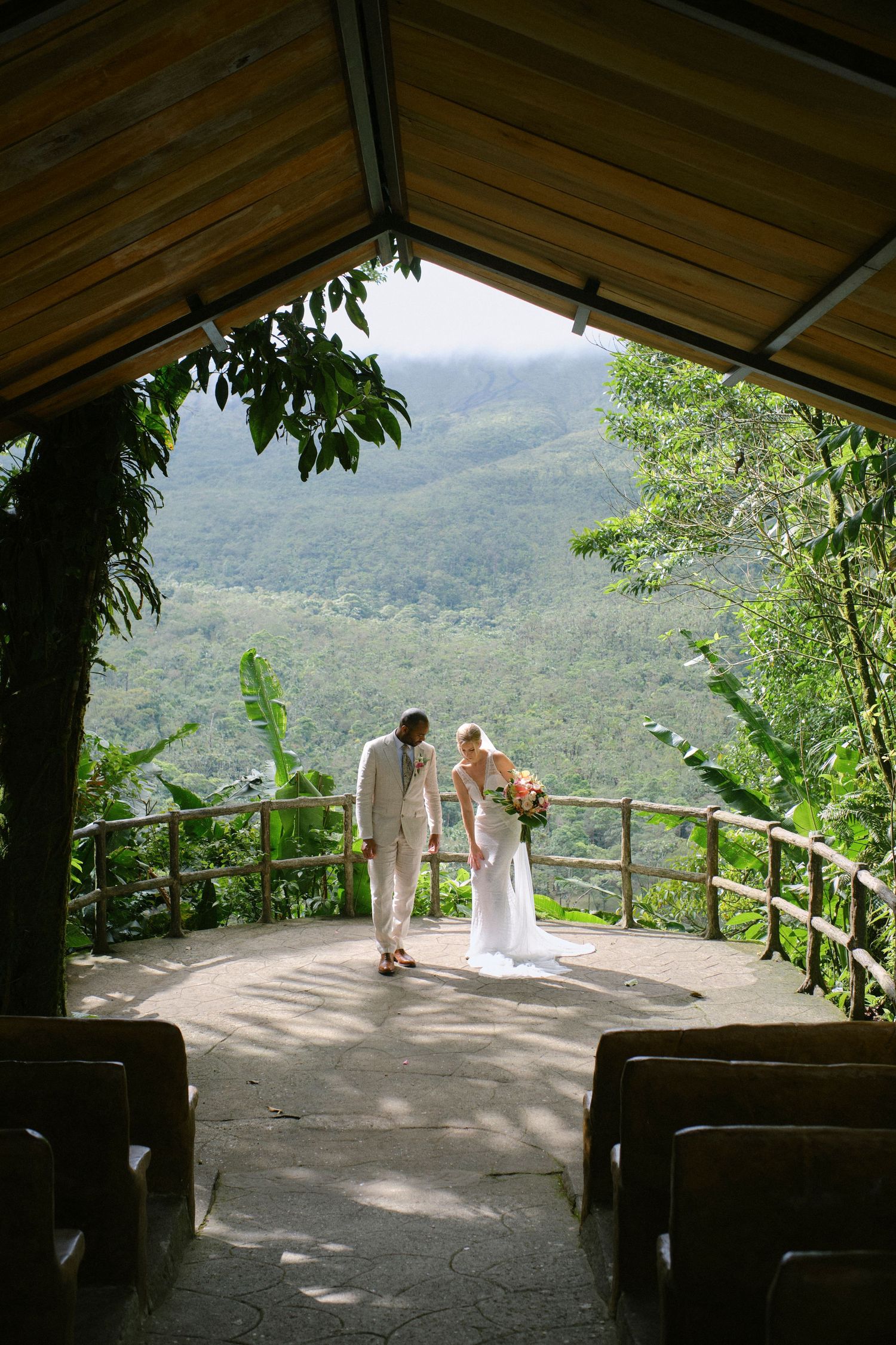 Bride and groom stand at an open-air chapel altar overlooking lush green tropical mountains on their wedding day.
