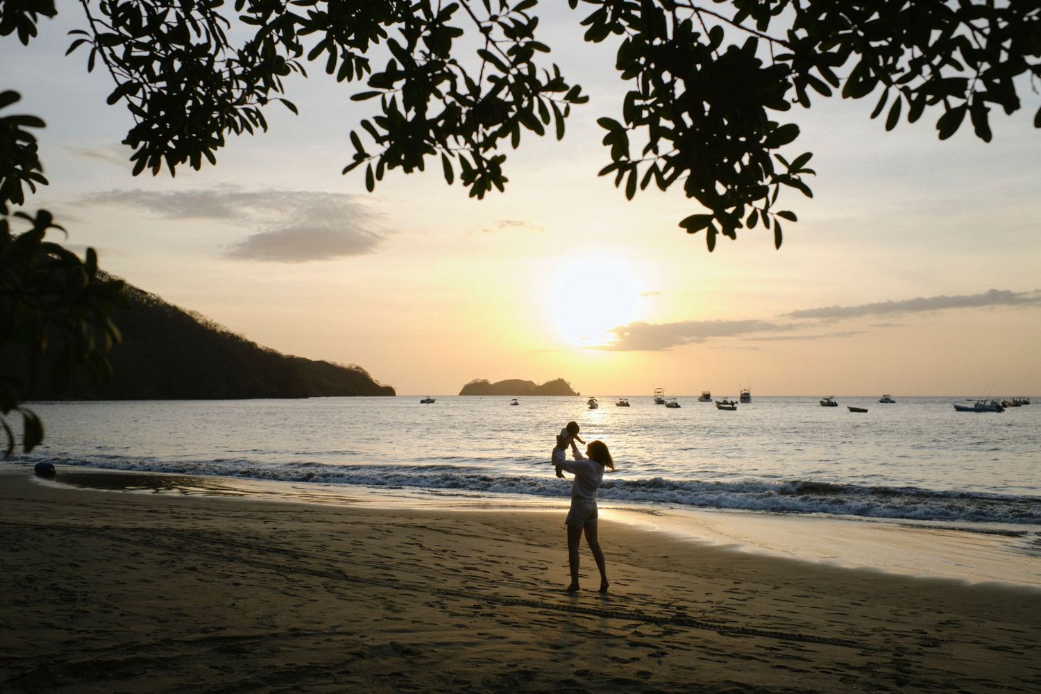 Silhouette of person holding child on tropical beach during stunning golden sunset over calm ocean waters.