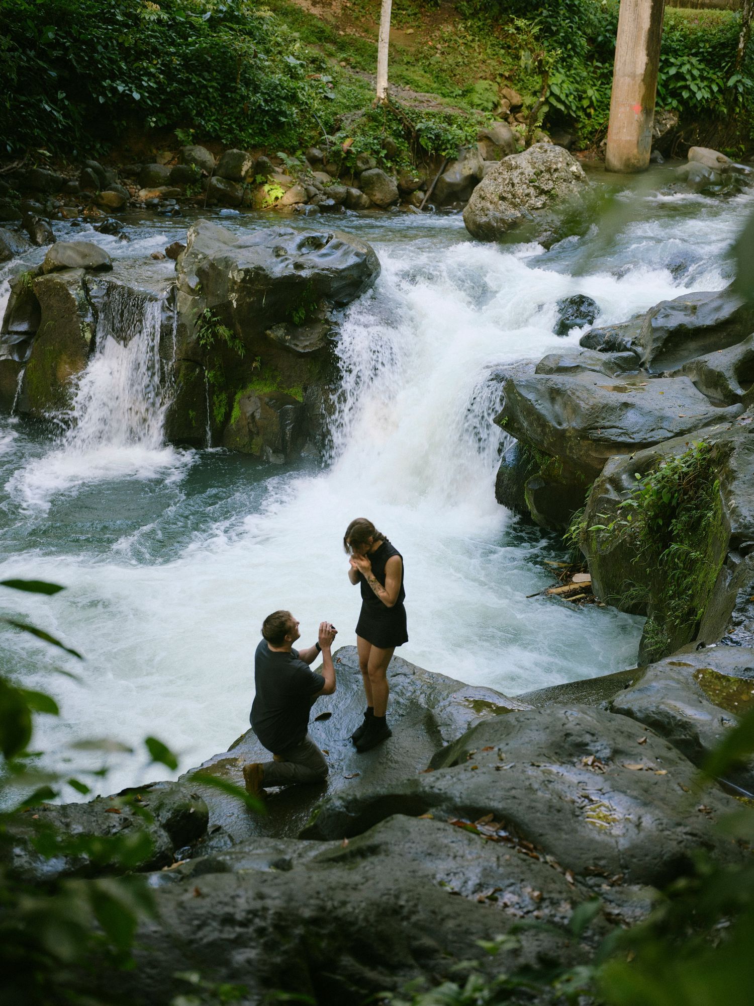 Man proposes on one knee to woman on rocks beside rushing waterfall in tropical rainforest.
