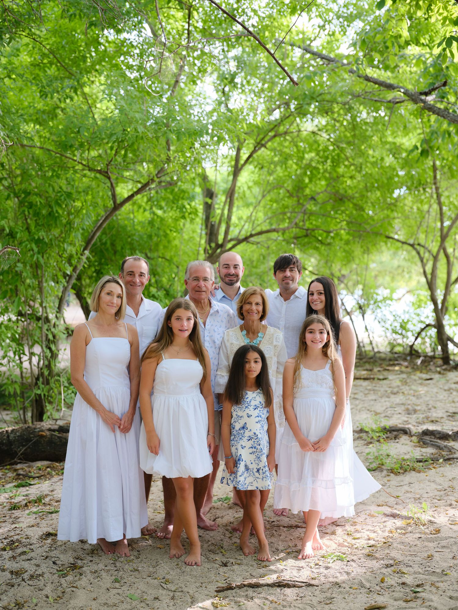 Large extended family in white dresses posing together on a sandy beach under shaded green trees.