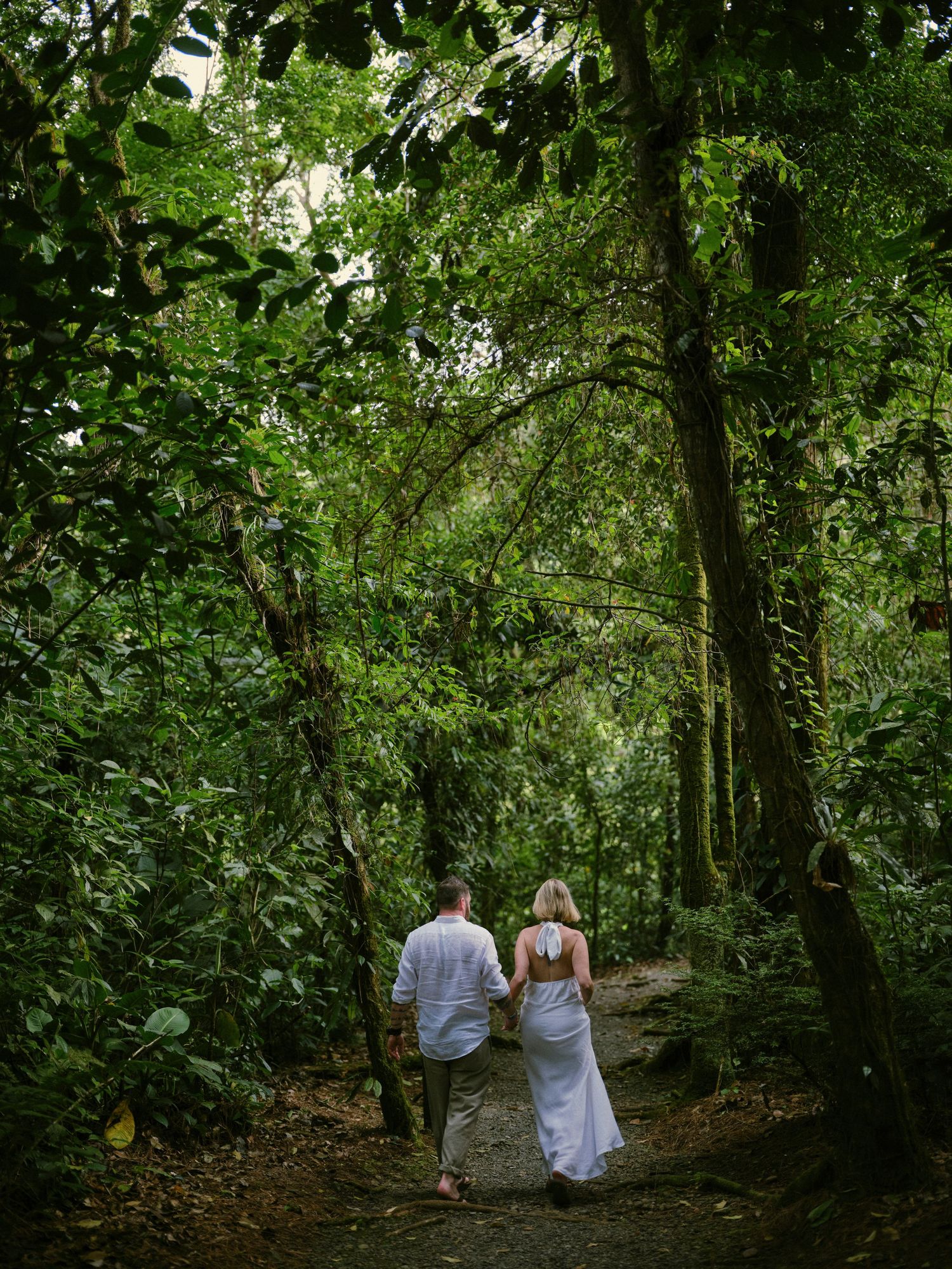 Bride and groom walking hand-in-hand along a shaded path through a dense tropical jungle forest.