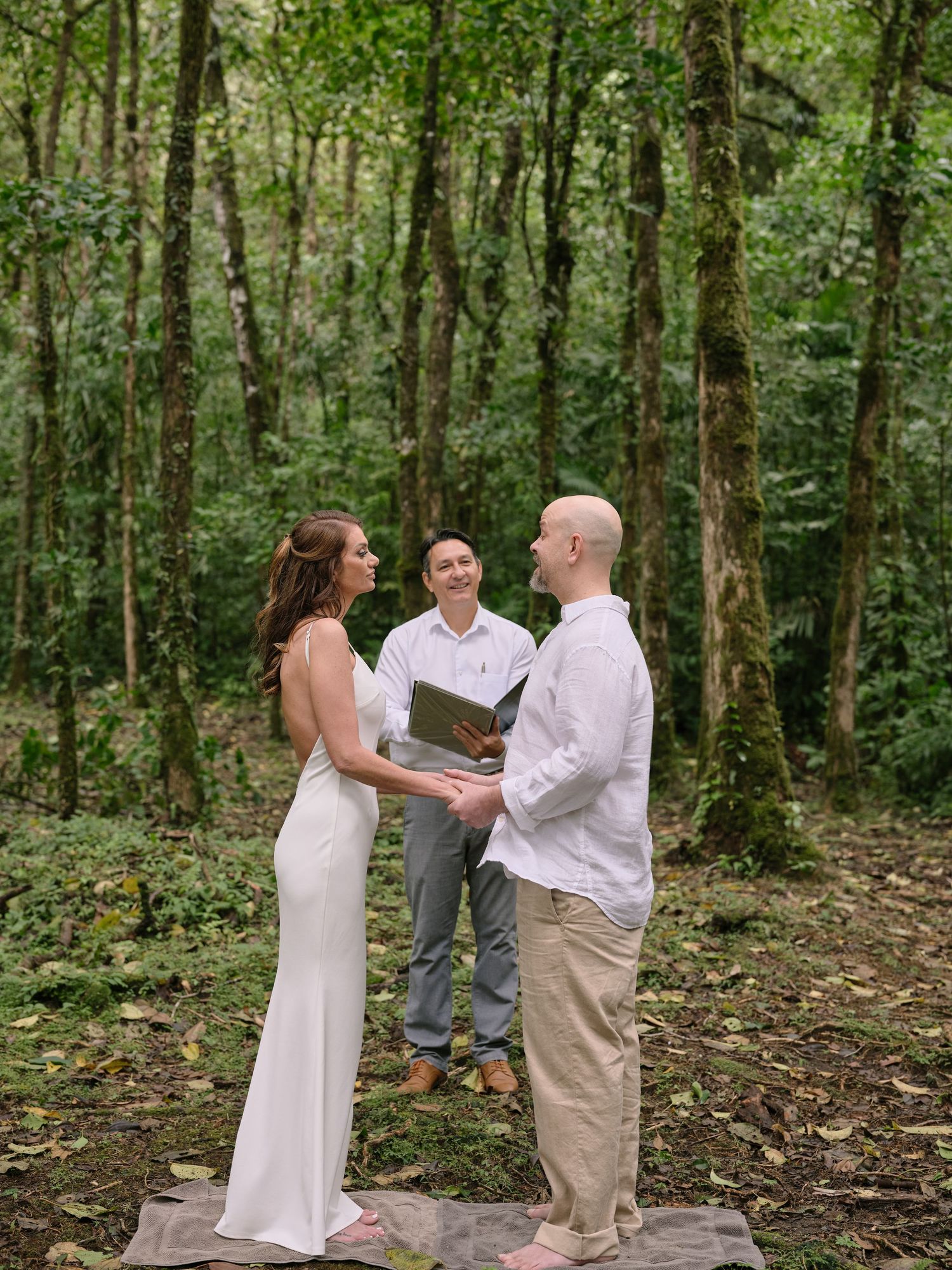Bride and groom exchanging vows in a lush forest ceremony with smiling officiant holding a book.