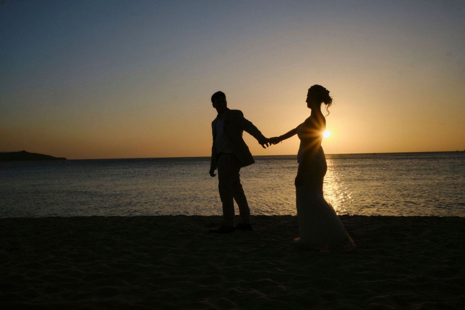 Silhouette of bride and groom holding hands on the beach during a golden sunset.