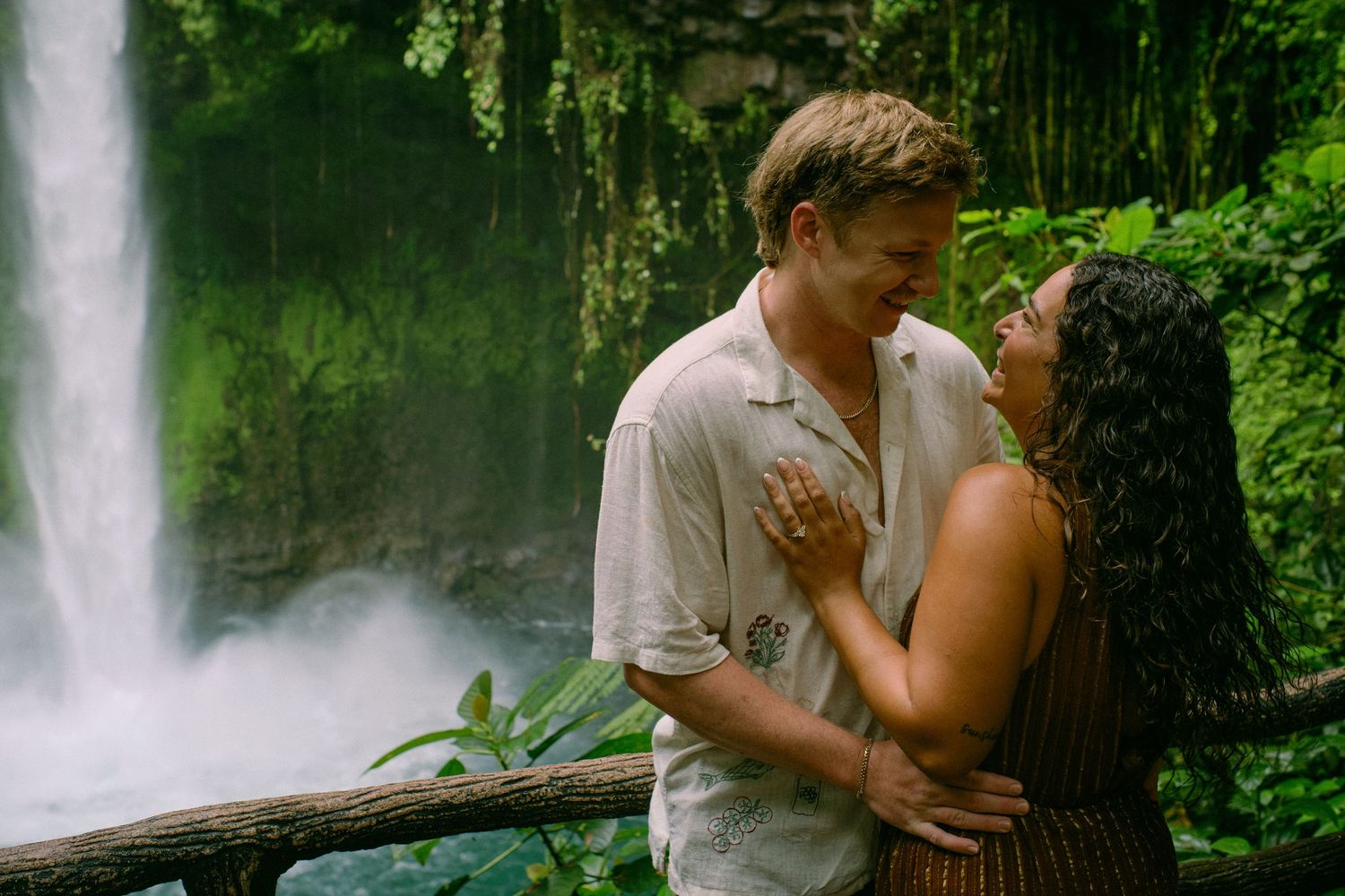Happy couple embracing and smiling at each other in front of a stunning tropical waterfall in lush jungle.