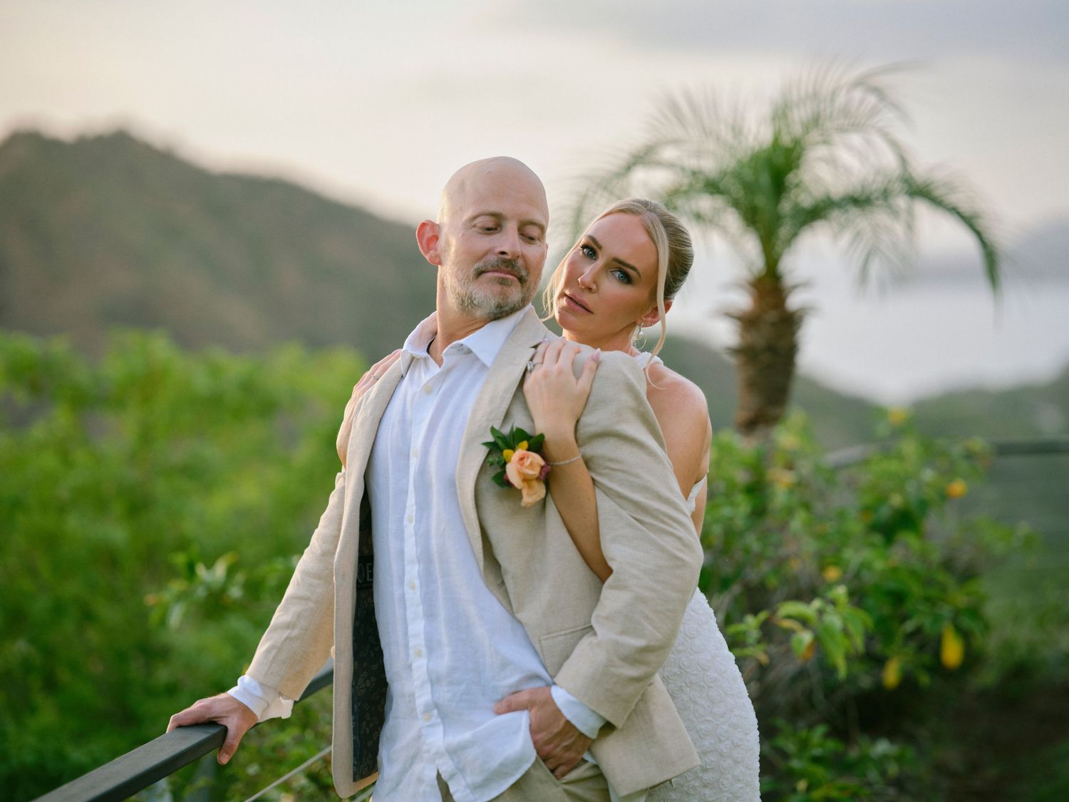 Couple in beige wedding attire poses on a tropical overlook with lush green mountains and palm trees at sunset.