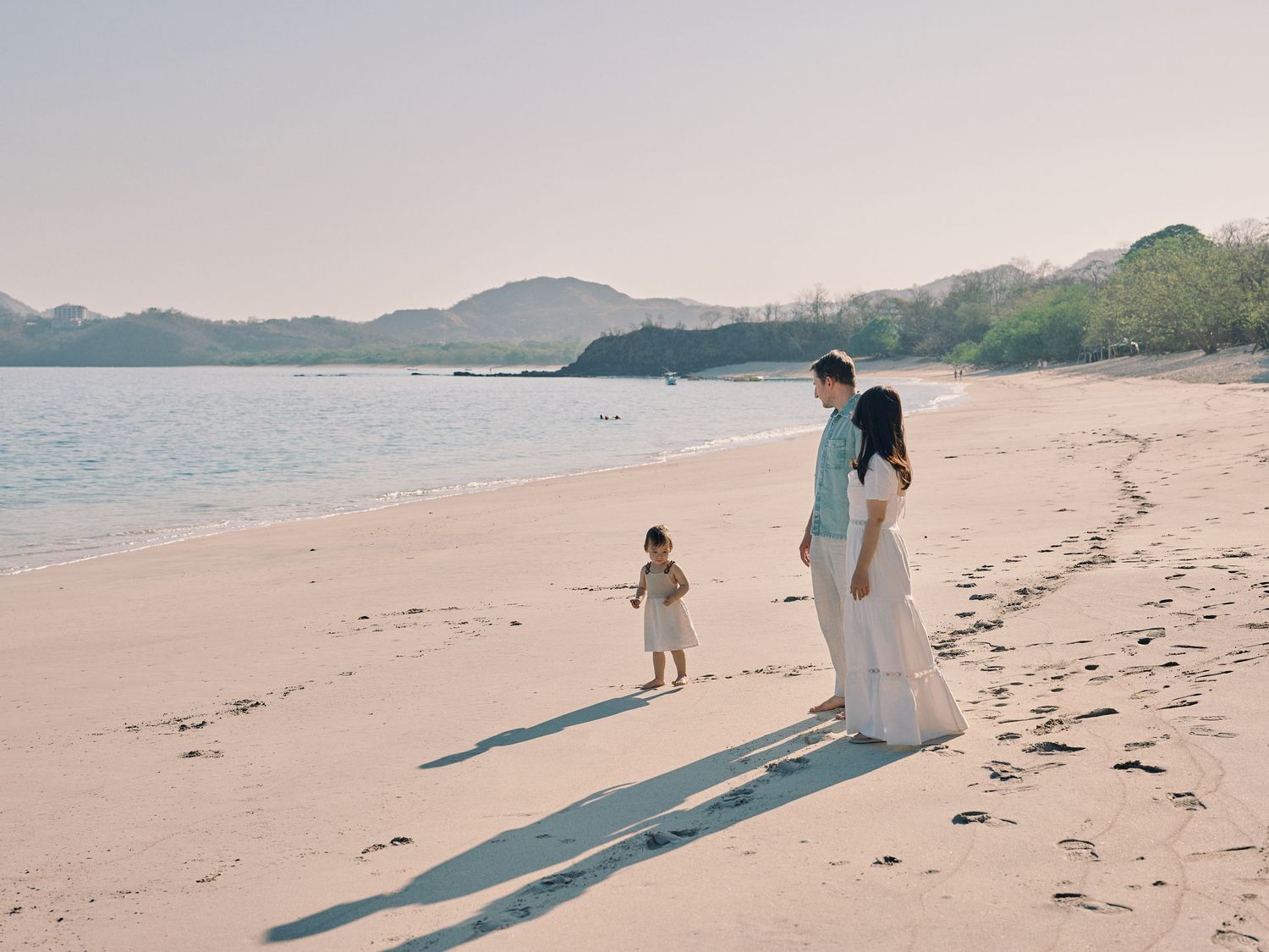 Parents walking with their young toddler along a serene sandy tropical beach at golden hour.