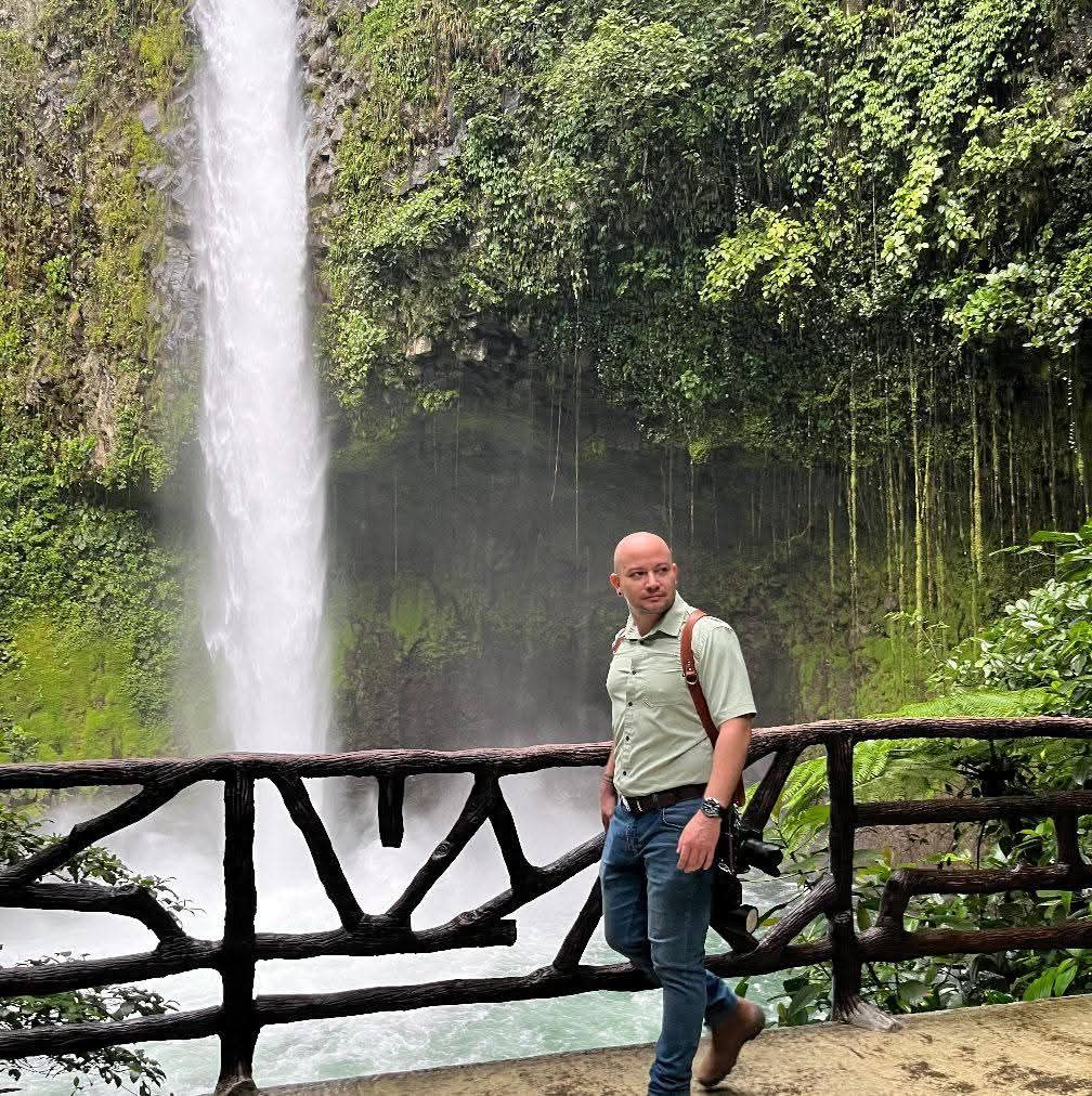 Man standing by rustic wooden fence with stunning tropical waterfall and lush green cliffs in background.