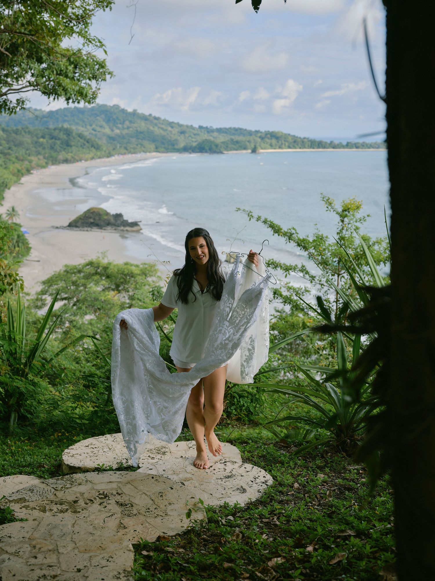 Bride in white lace dress twirling on stone overlook above tropical beach coastline.