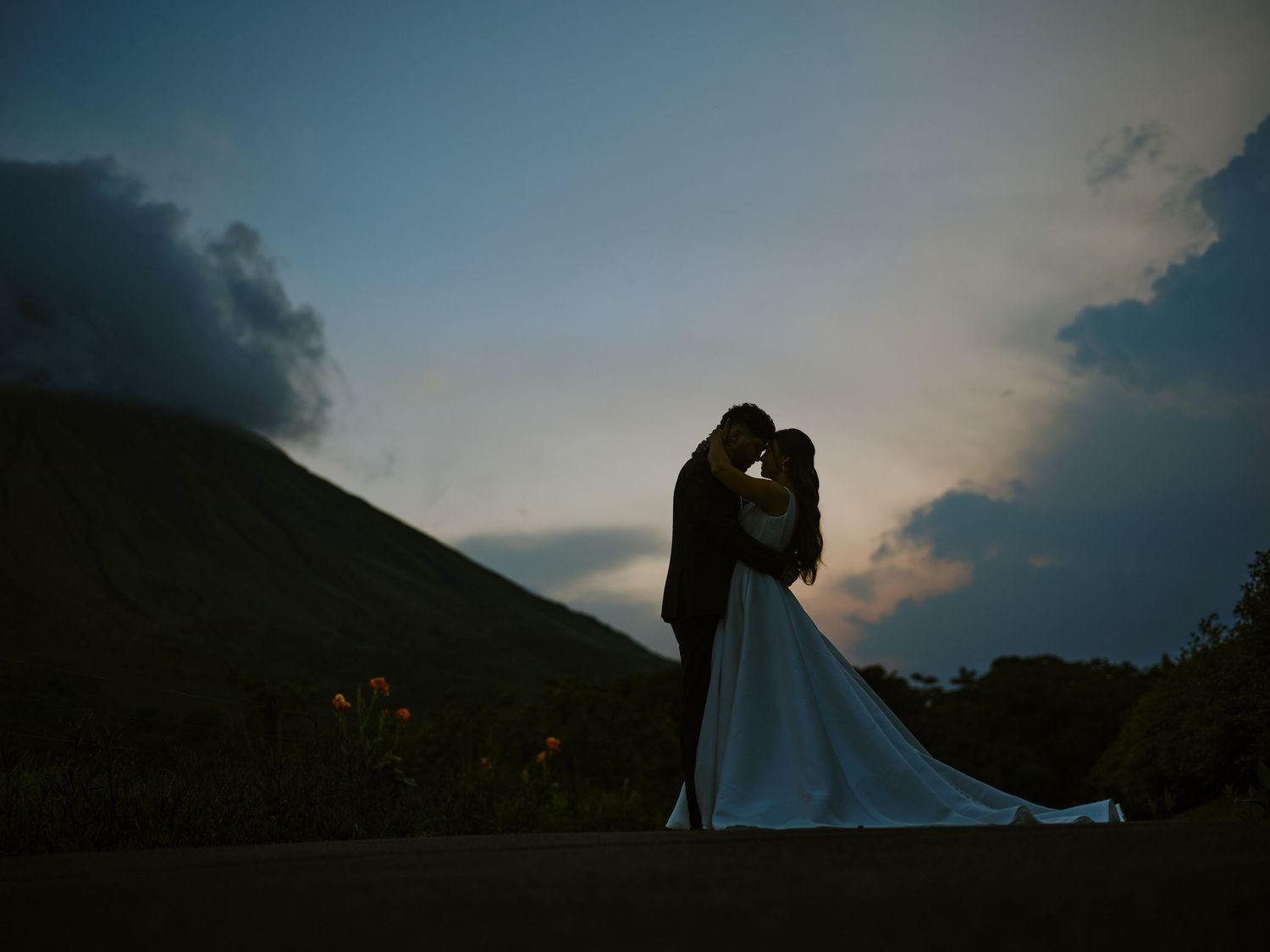 Romantic silhouette of bride and groom embracing at dusk with mountain and colorful sky behind them.