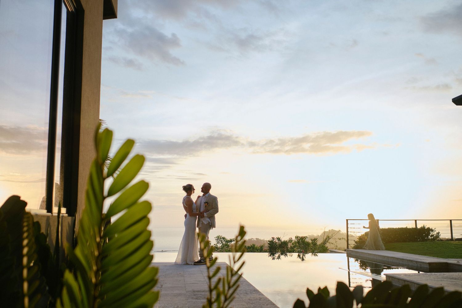 Silhouetted bride and groom stand by an infinity pool under a dramatic golden sunset sky at a resort.
