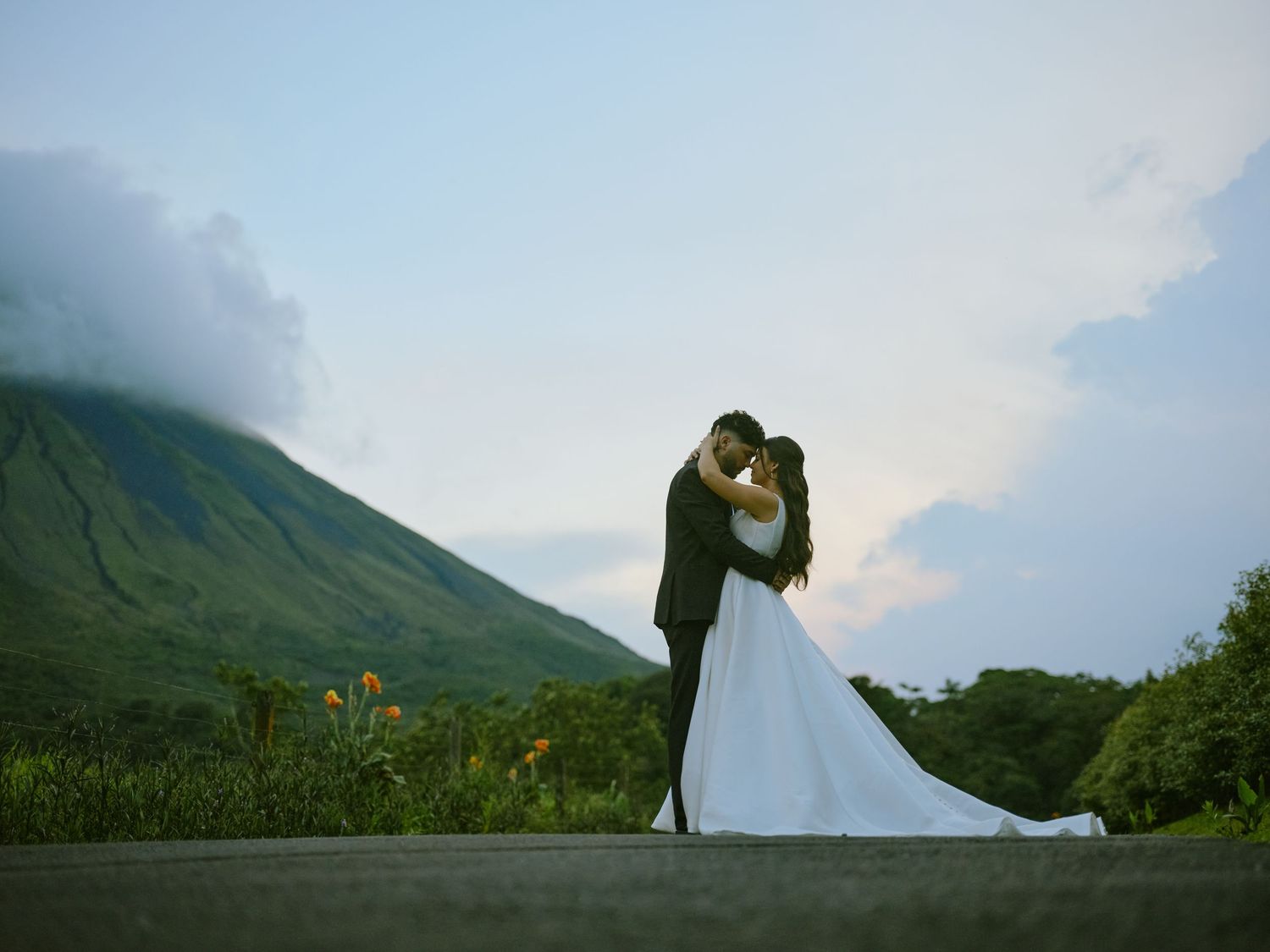 Bride and groom embracing with lush green volcano and dramatic clouds in background at outdoor wedding.