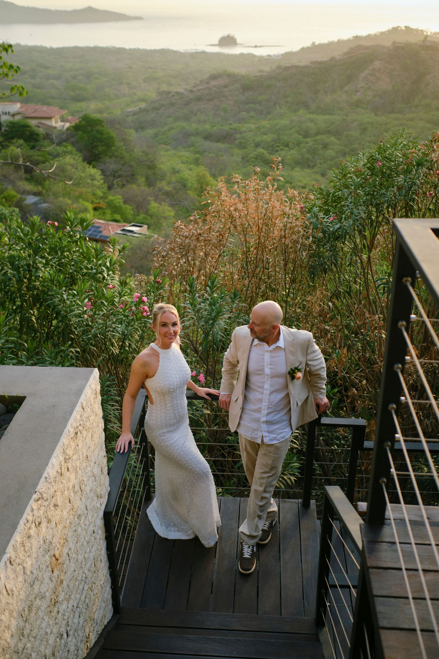 Bride and groom walk up outdoor stairs with a scenic tropical coastal valley view at sunset behind them.