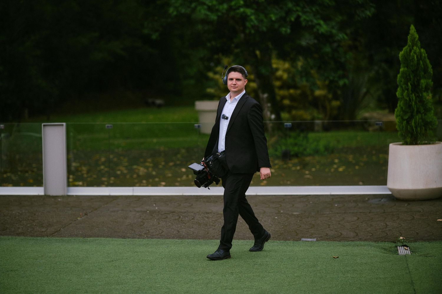 Man in black suit walking outdoors on green turf holding camera equipment with lush garden background.