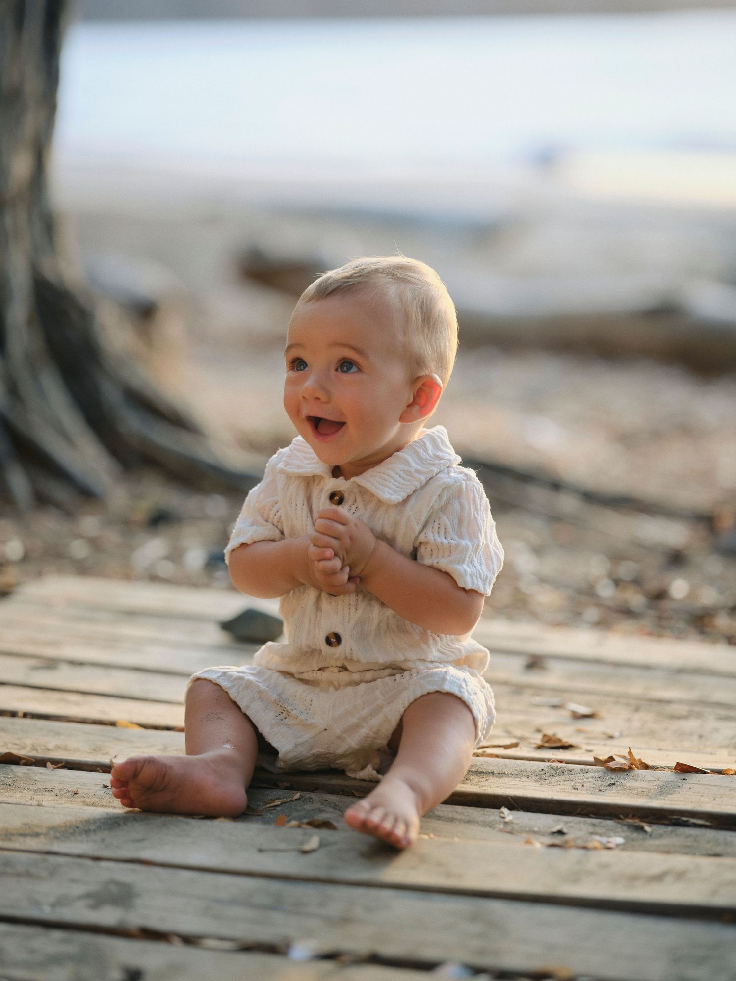 Adorable smiling baby in cream outfit sitting on wooden boards near a tree on a sandy beach.