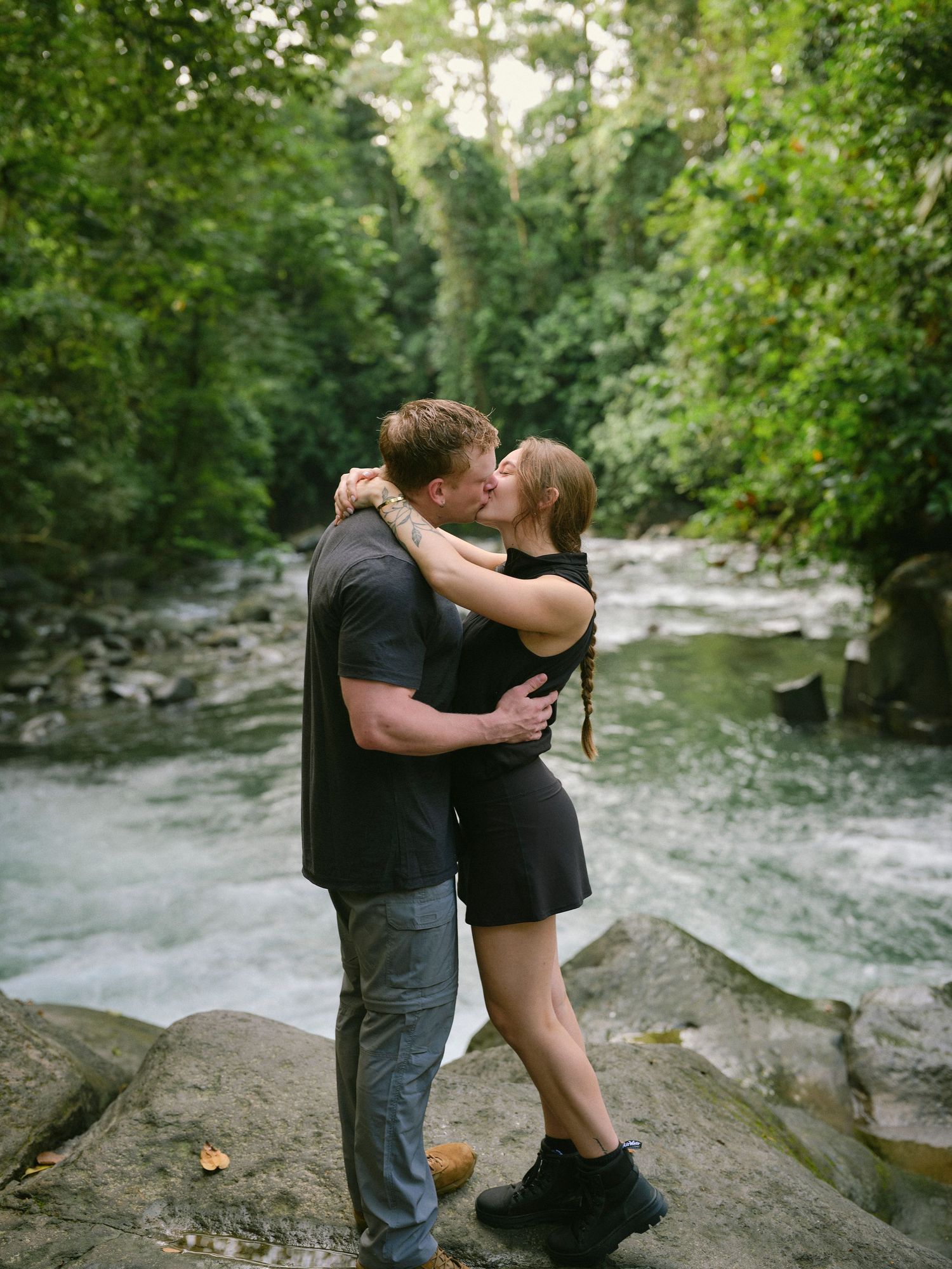 Couple shares passionate kiss on rocky riverbank with flowing stream and green forest behind them.