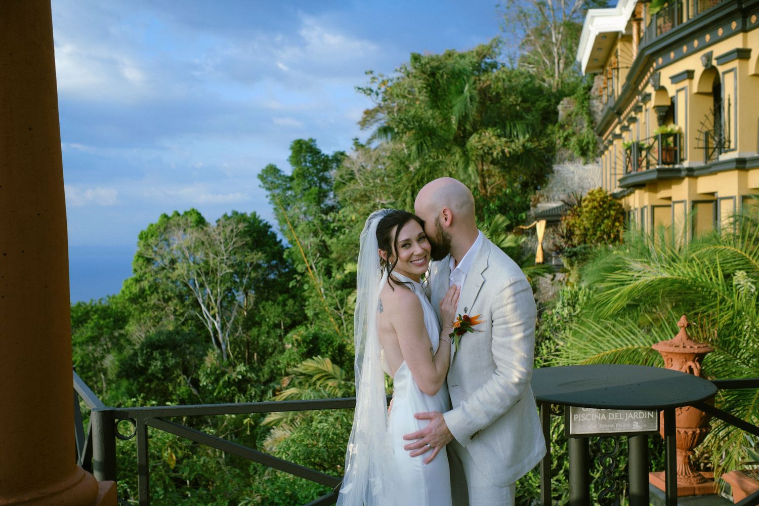 Bride and groom share a romantic kiss on a balcony overlooking lush tropical scenery.