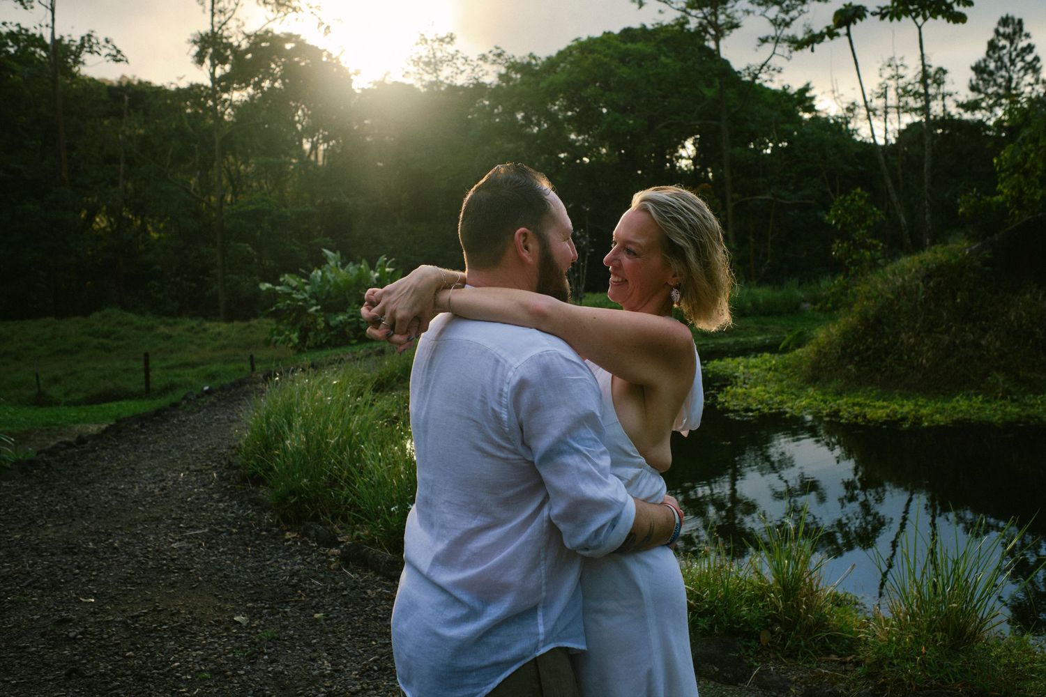 Couple embracing and dancing near a pond at golden sunset surrounded by tropical greenery.