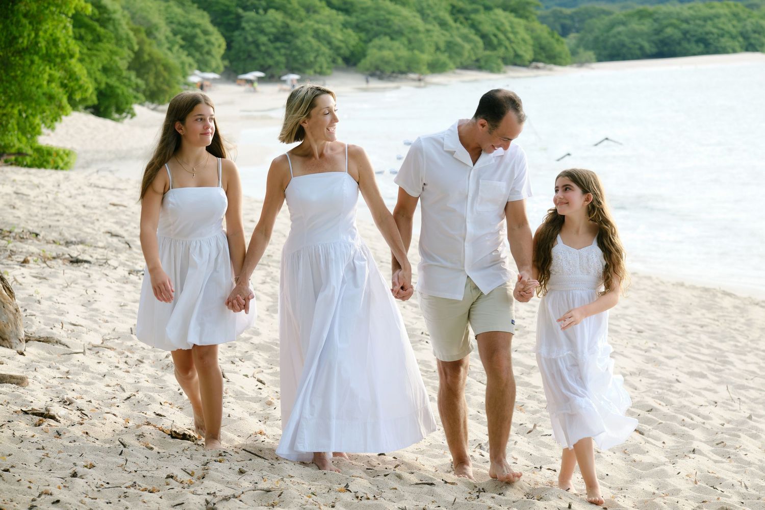 Family of four in white outfits walking hand in hand along a sunny tropical beach with green trees.
