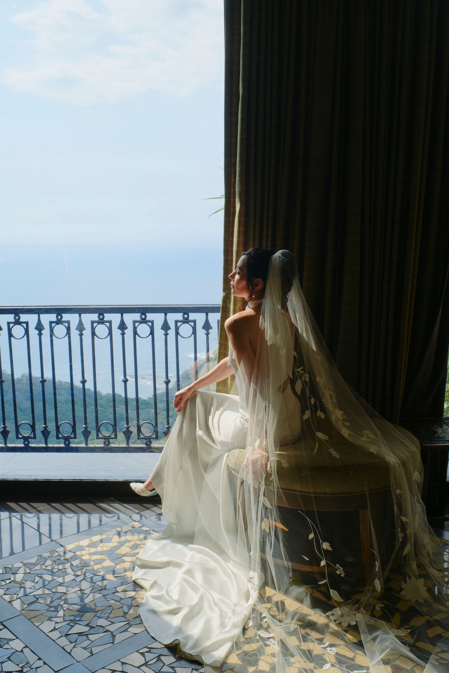 Bride in white gown with floral veil sits by a sunlit balcony overlooking a scenic landscape.